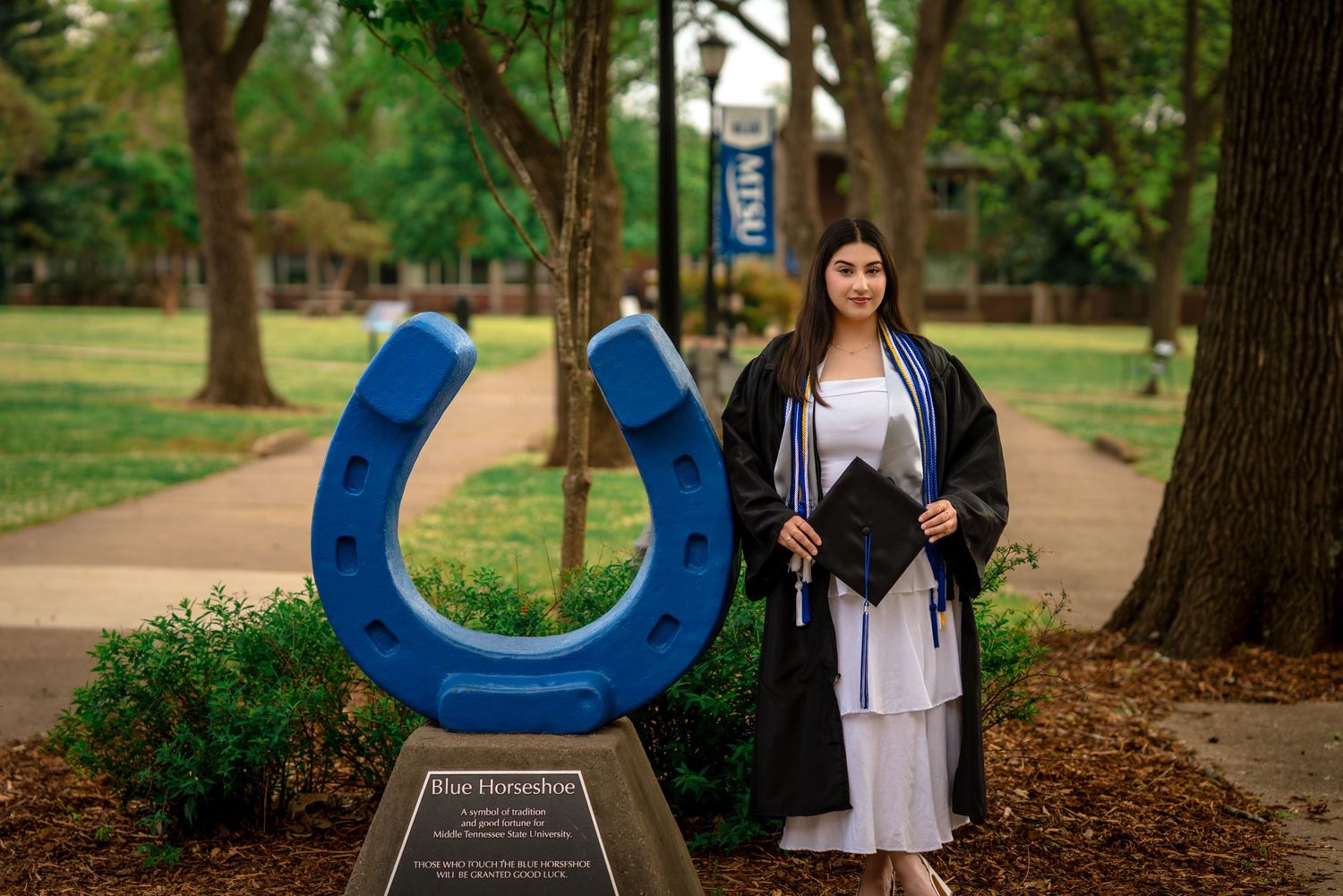 College graduate in cap and gown poses beside a blue horseshoe sculpture on a tree-lined campus walkway.