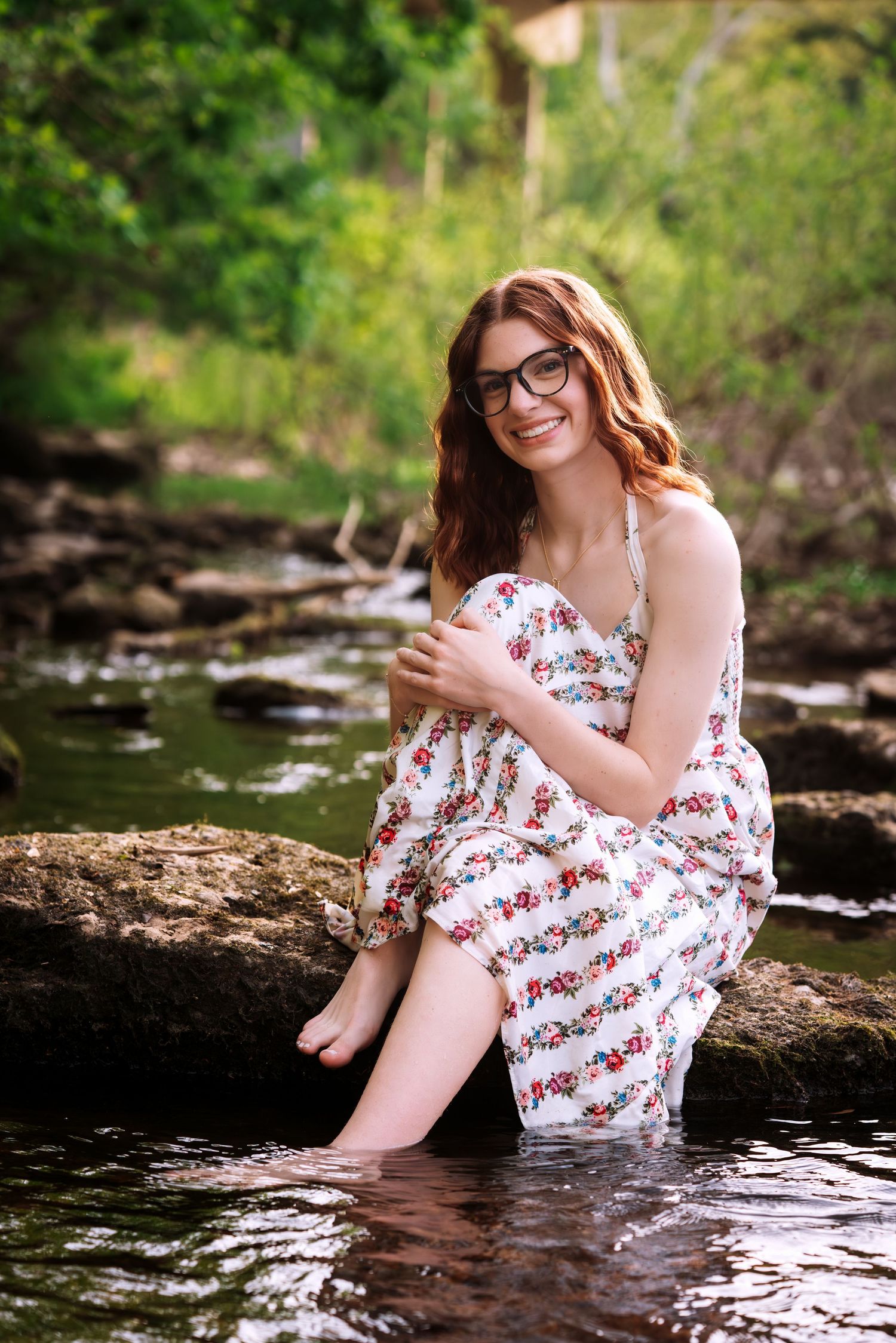 Young woman in floral dress sitting on rocks in a stream, smiling, wearing glasses, feet in water.
