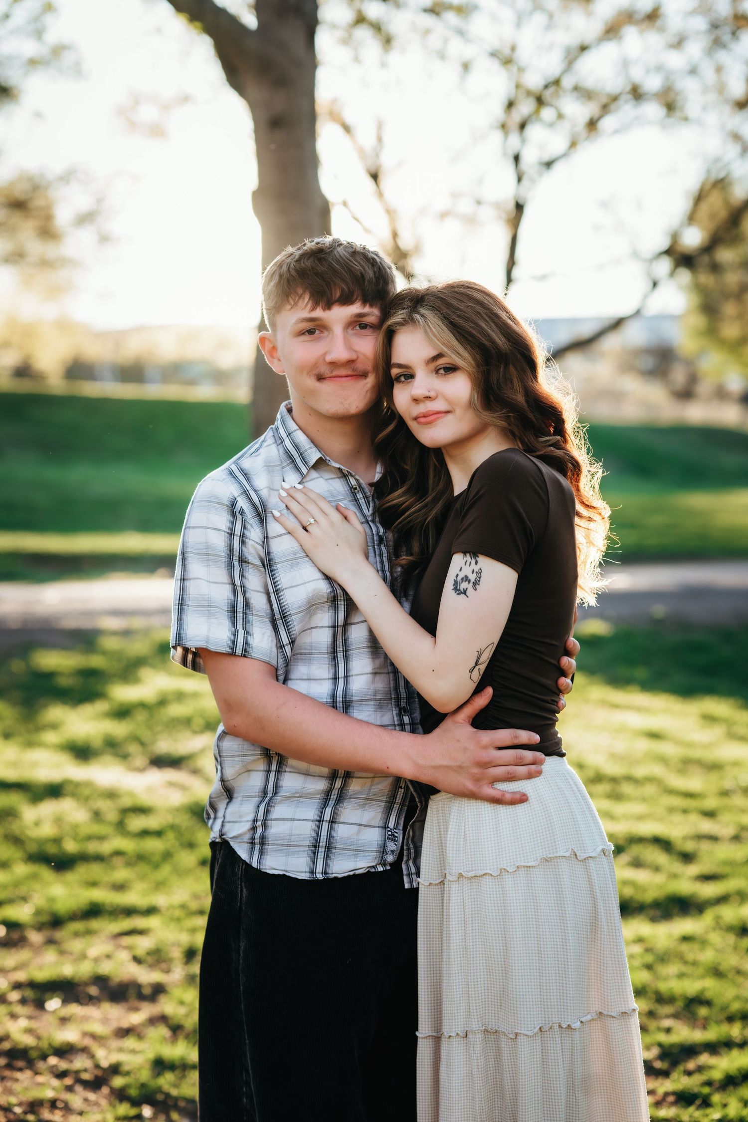 A smiling couple embraces in a sunlit park, the woman showing an engagement ring while leaning against her partner.