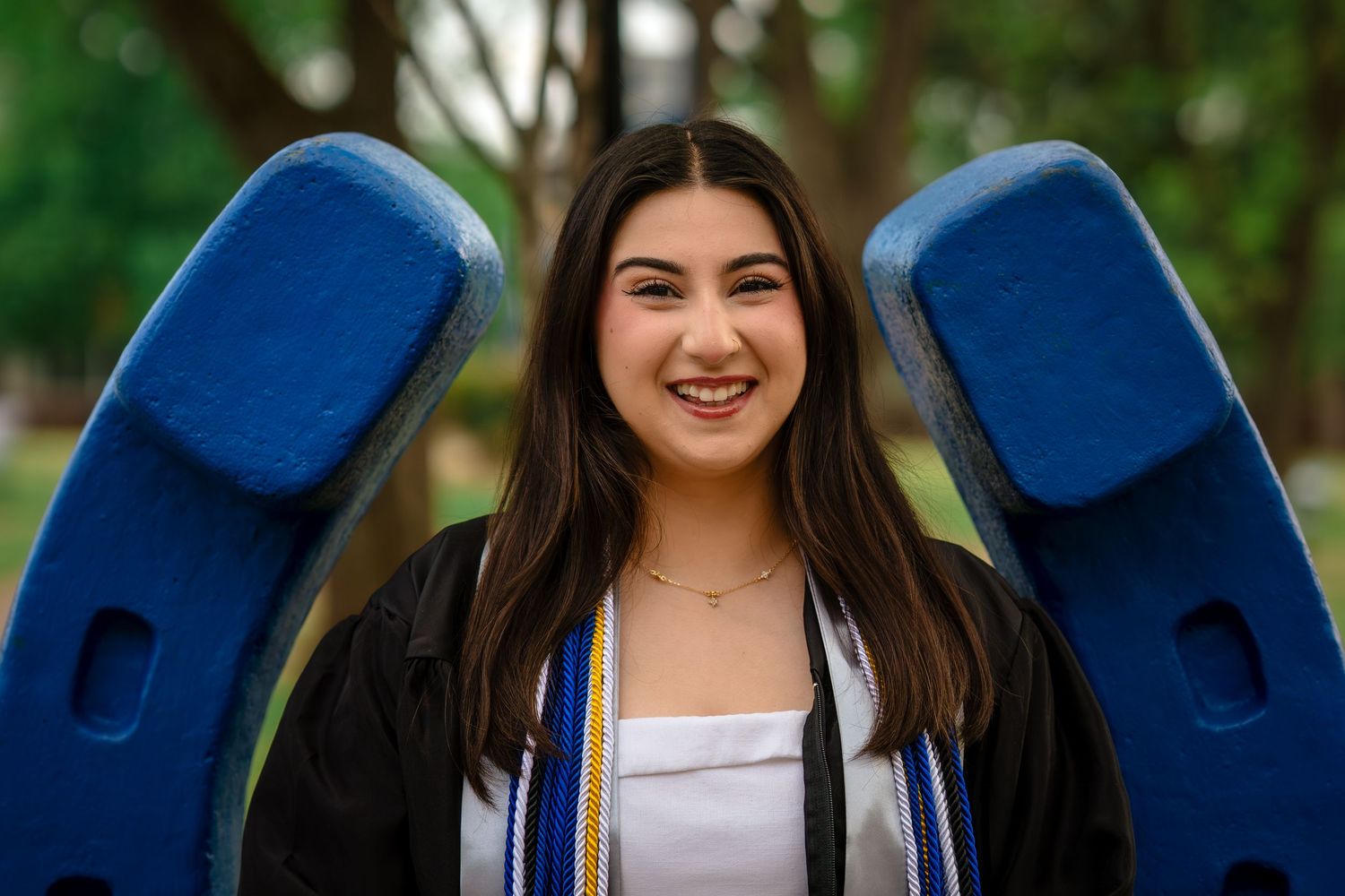 Smiling female graduate in cap and gown with honor cords poses outdoors near blue university letter sculpture.