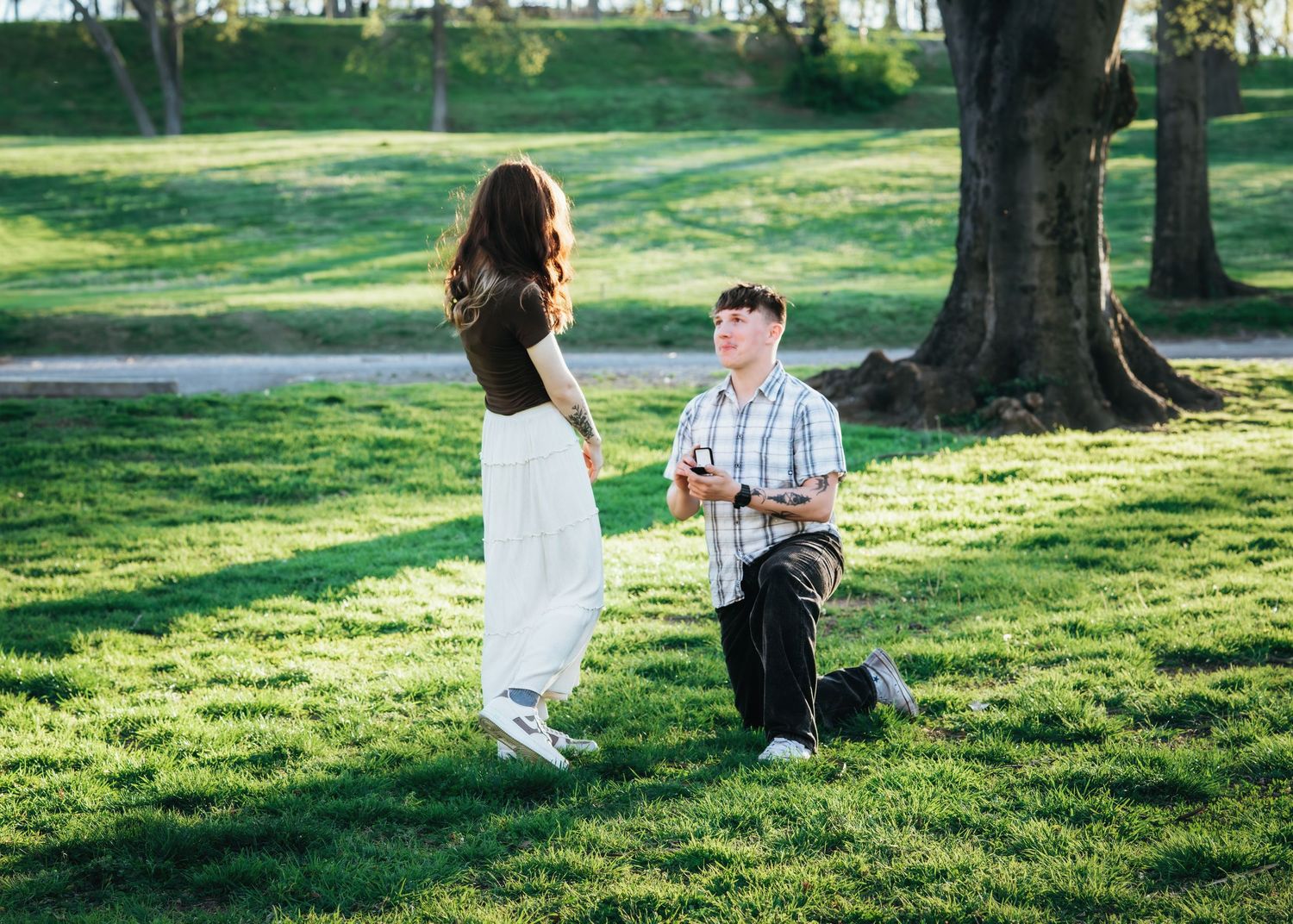 Man kneeling on one knee proposing to woman on green grass in sunlit park.