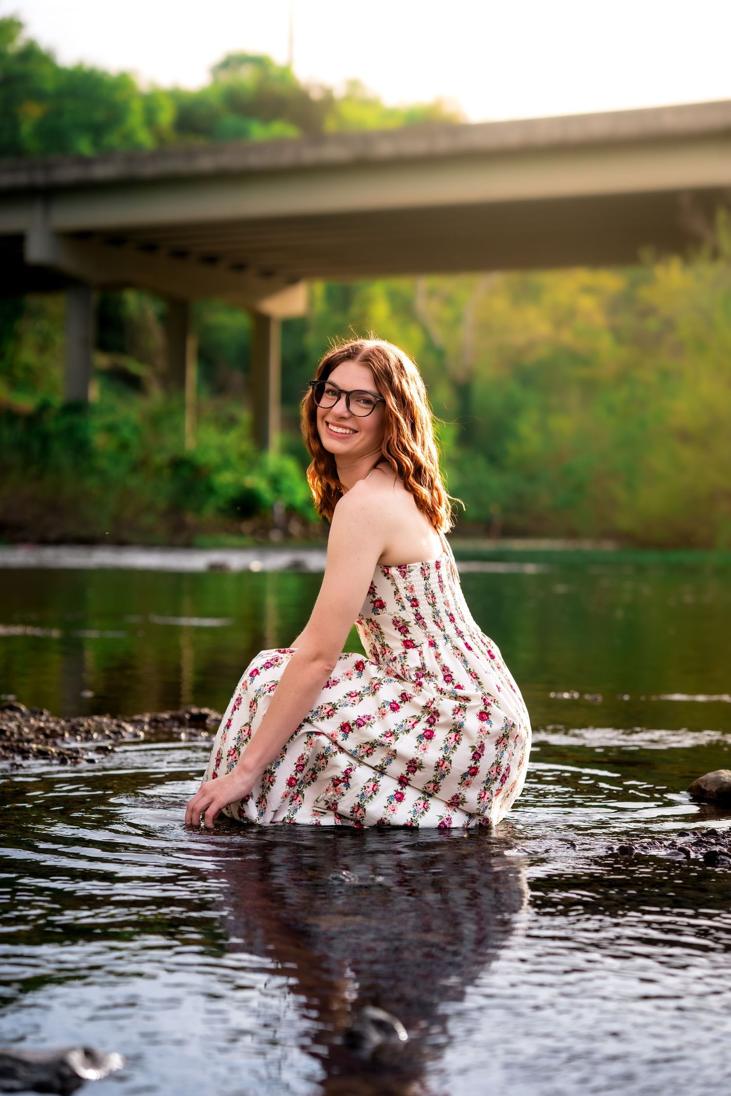 Smiling woman in floral dress crouching in a shallow river with a concrete bridge behind her.