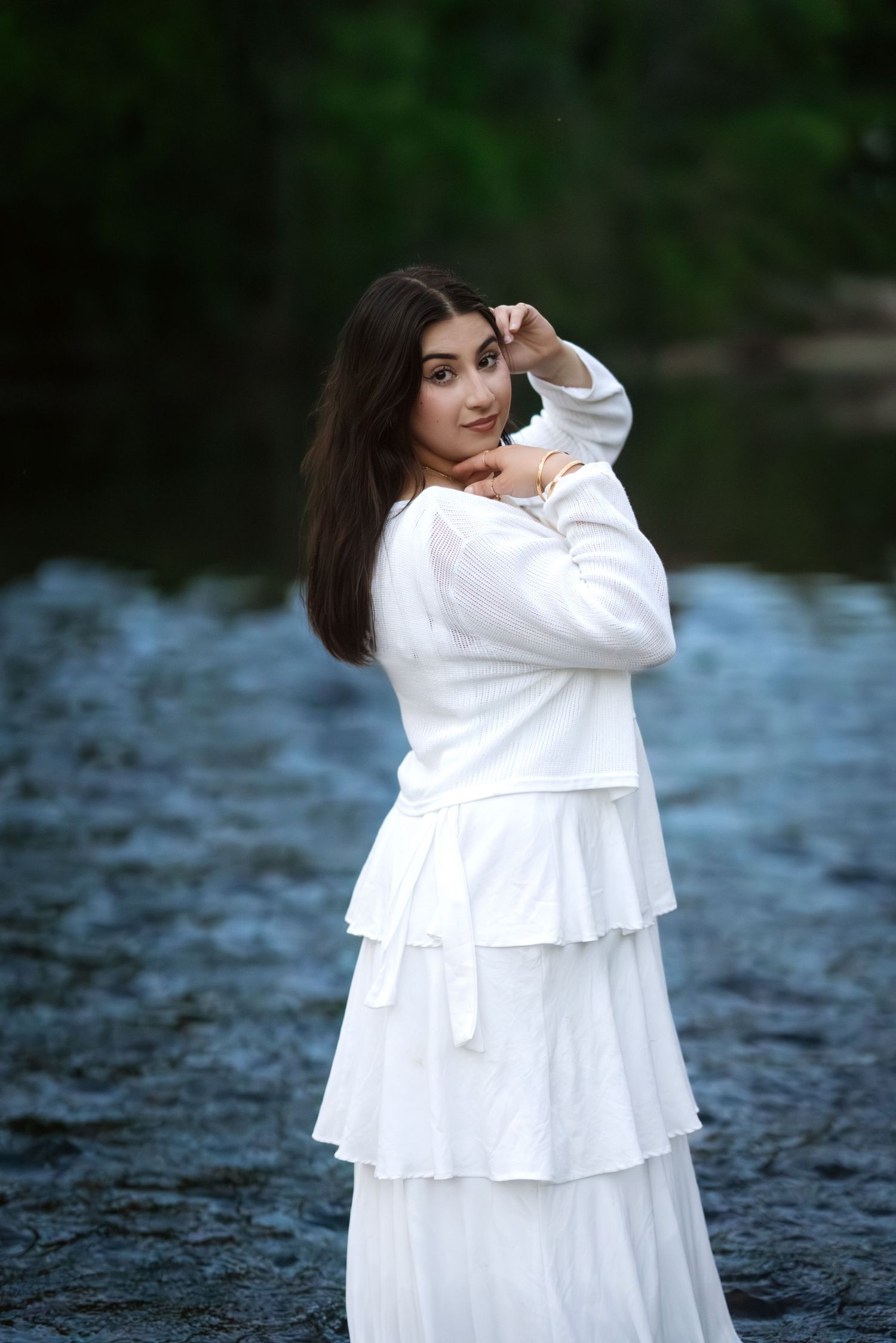 Brunette woman in elegant white layered dress posing gracefully by a serene river.