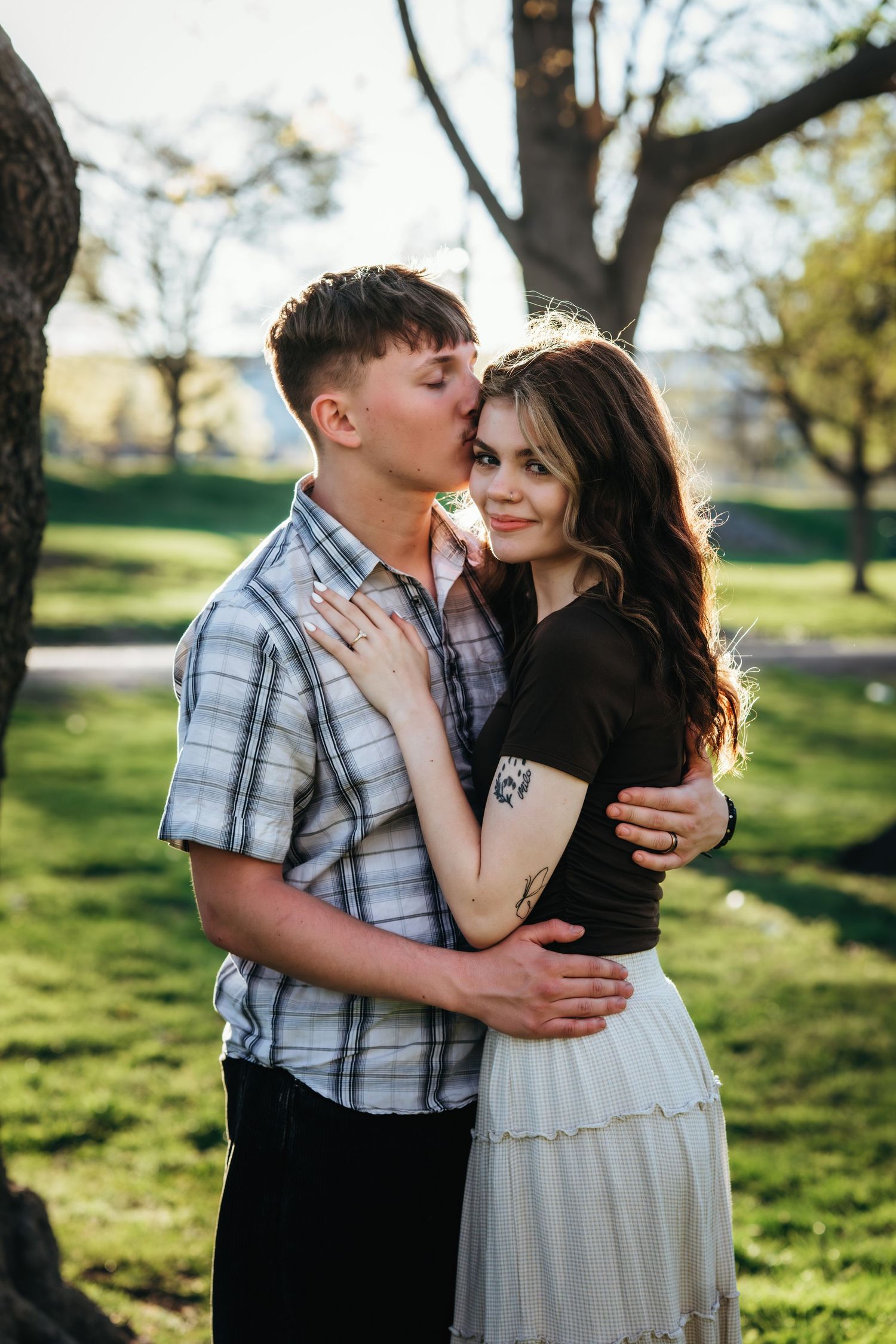 Young couple embracing in a green park at golden hour, man kissing woman on the cheek as she smiles.
