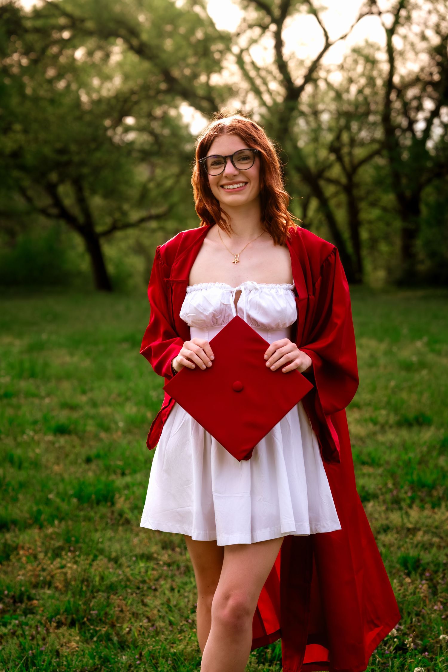 Smiling graduate in red gown and white dress holding her cap in a green park.