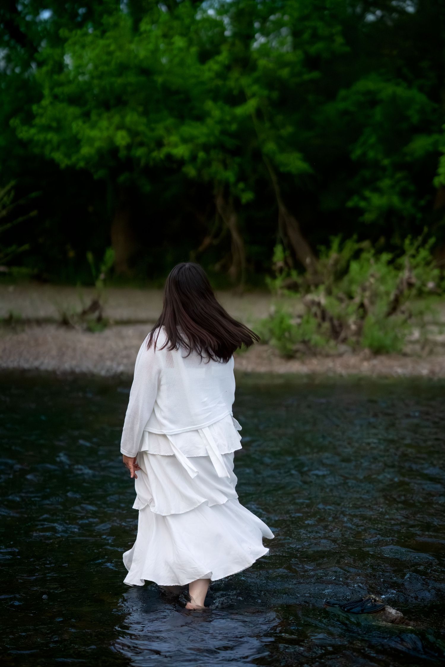 Woman in white tiered dress wading barefoot in calm river surrounded by green trees.