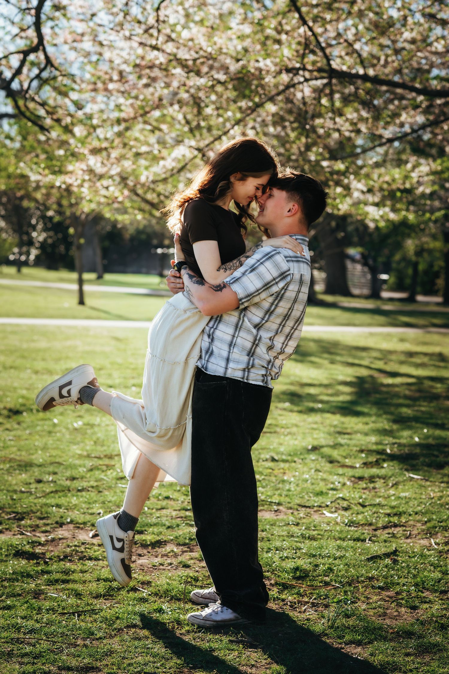 Romantic couple in a sunny park, man lifting woman in his arms beneath flowering cherry blossom trees.