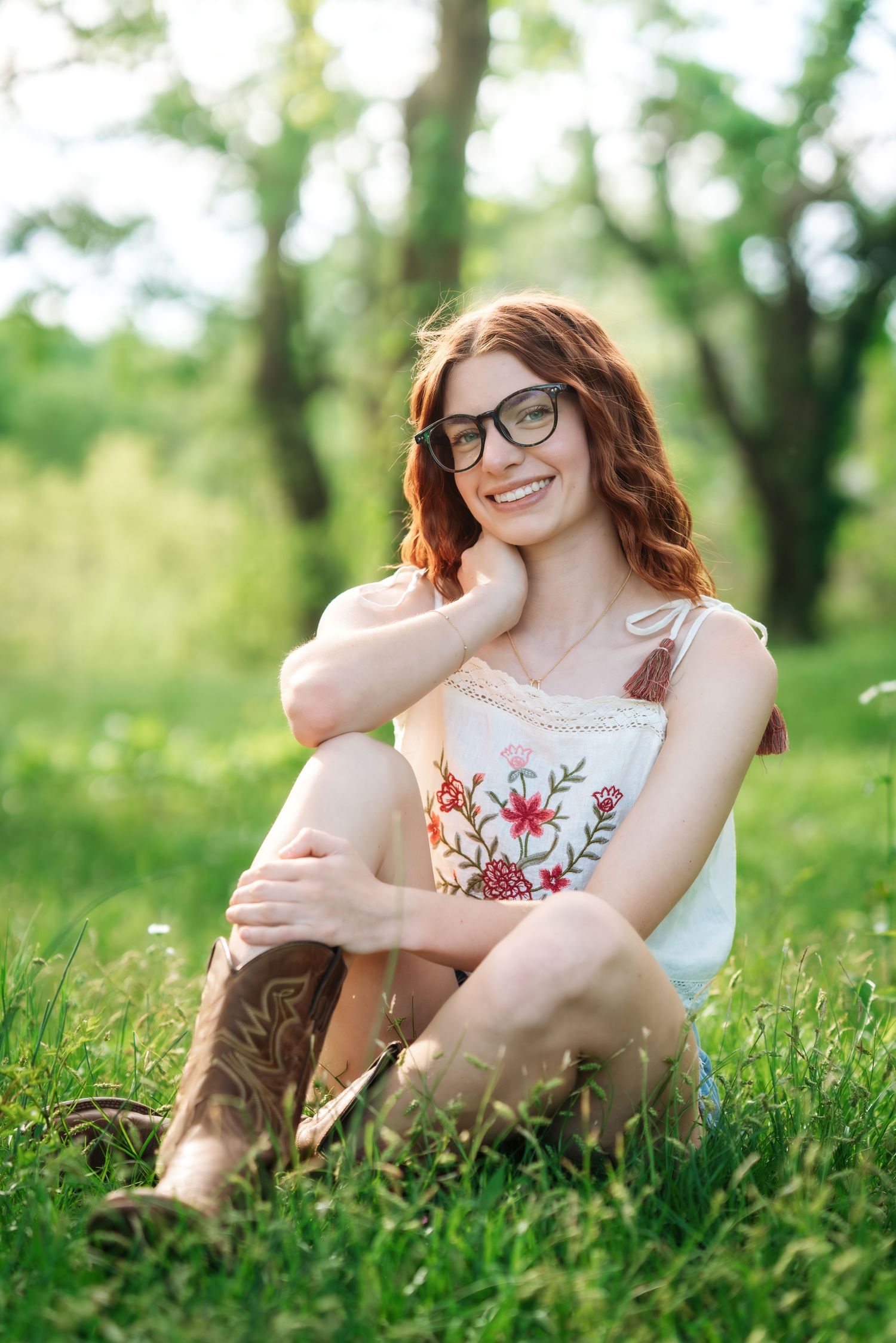 Happy redhead woman wearing glasses and cowboy boots sitting on green grass in a sunny park setting.