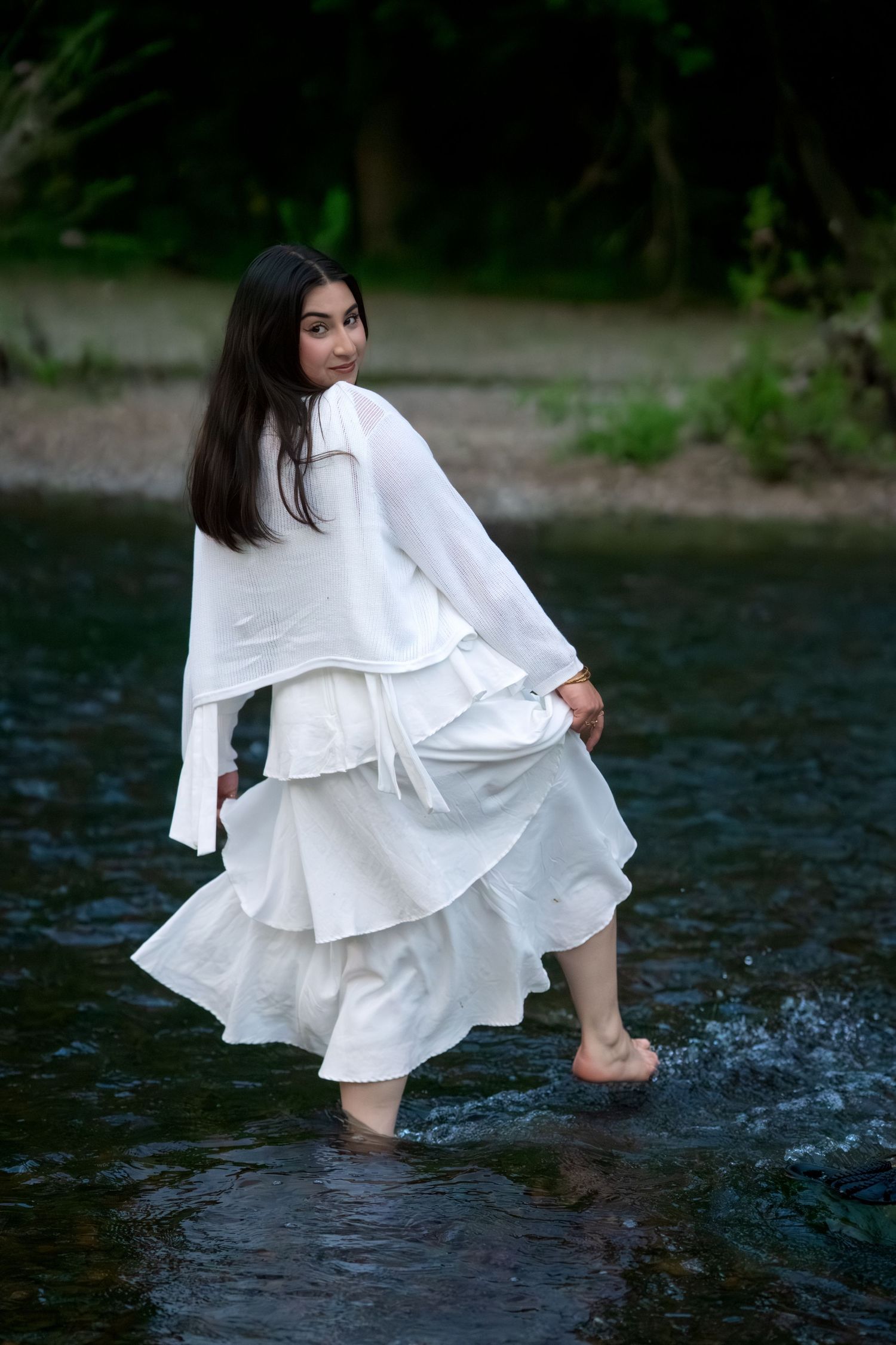 Woman in flowing white dress wades barefoot through a calm river surrounded by greenery.