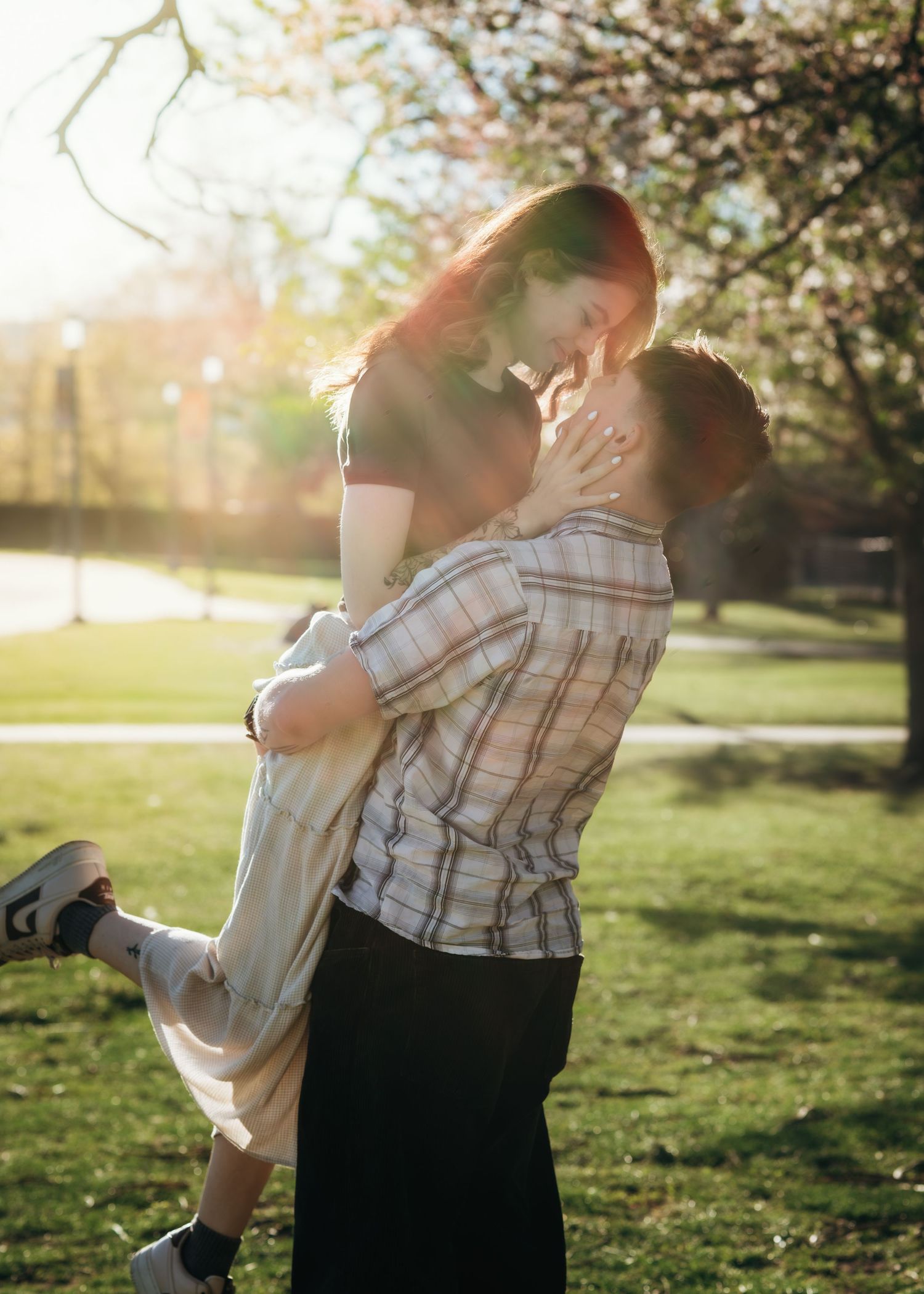 Man in plaid shirt lifts woman in sunlit park, sharing a romantic near-kiss moment outdoors.