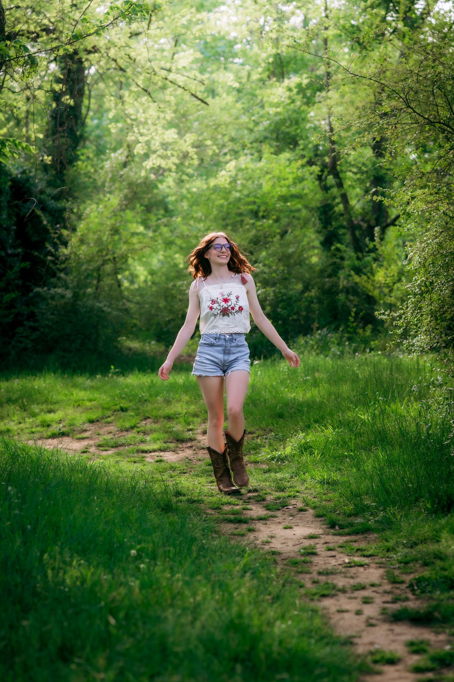 Young woman in denim shorts and cowboy boots walks along a sunlit forest path surrounded by lush green trees.