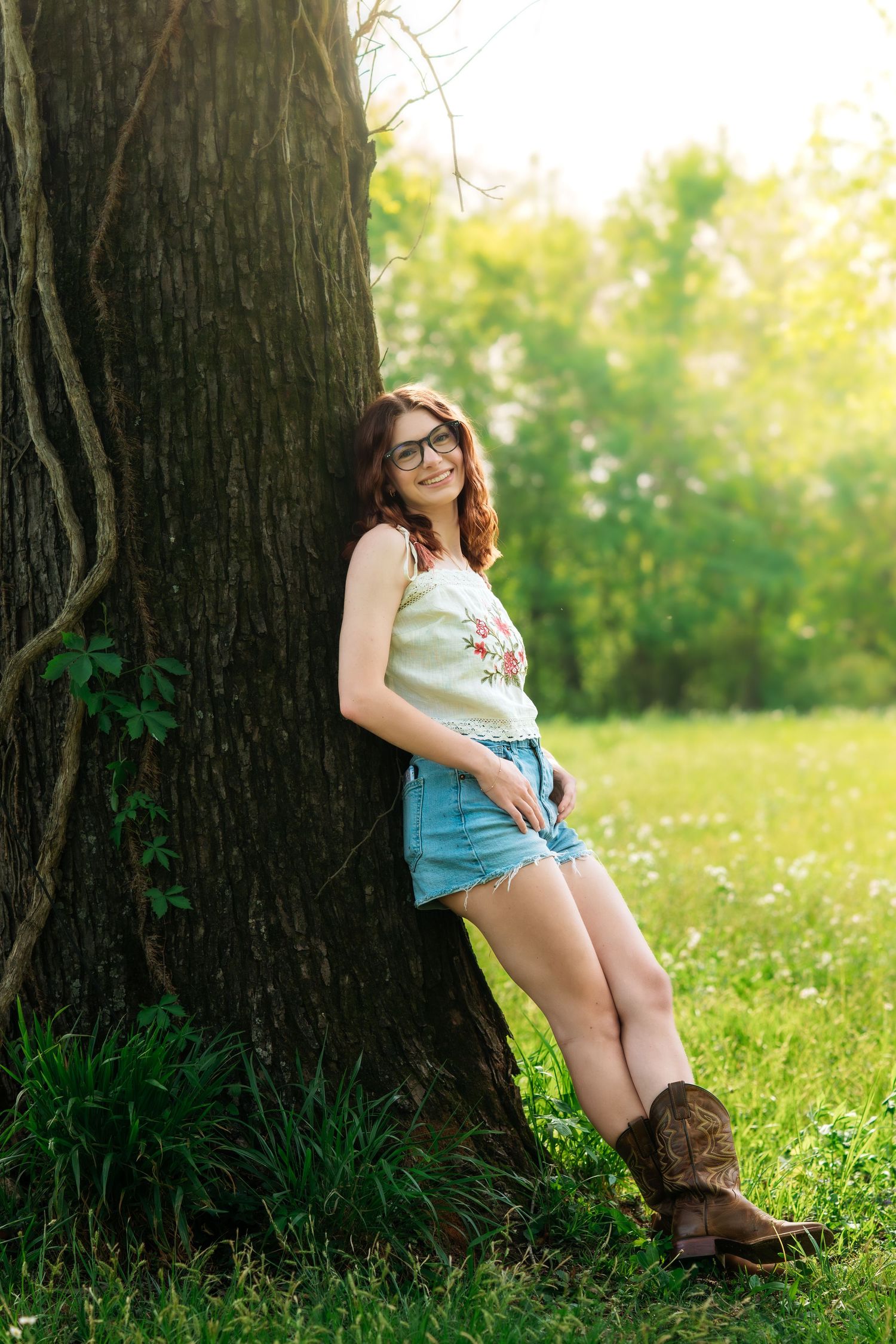 Young woman in denim shorts and floral top leaning against a large tree in a sunny green park.