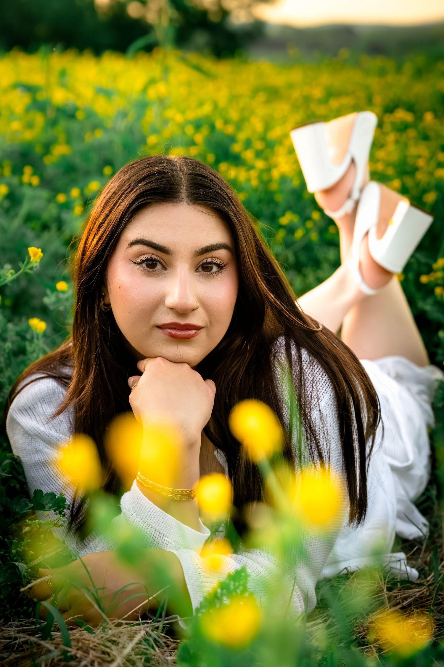 Young woman lying in a vibrant yellow wildflower field, resting chin on hand, wearing white outfit and heels.