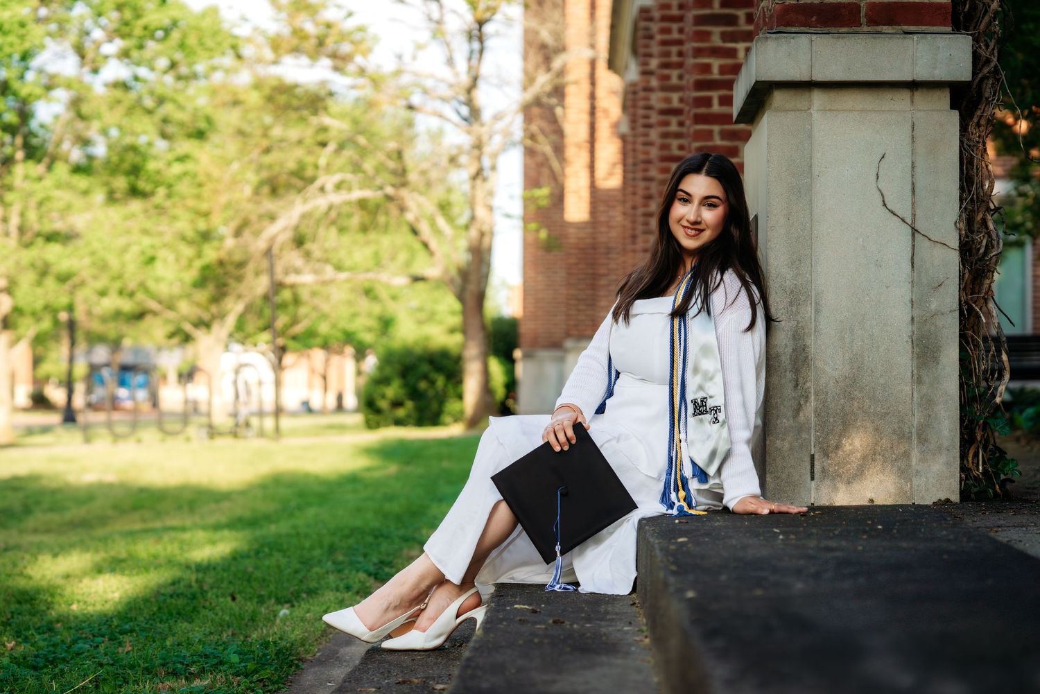 Young woman in white graduation attire holding cap, seated outdoors near brick building on campus.