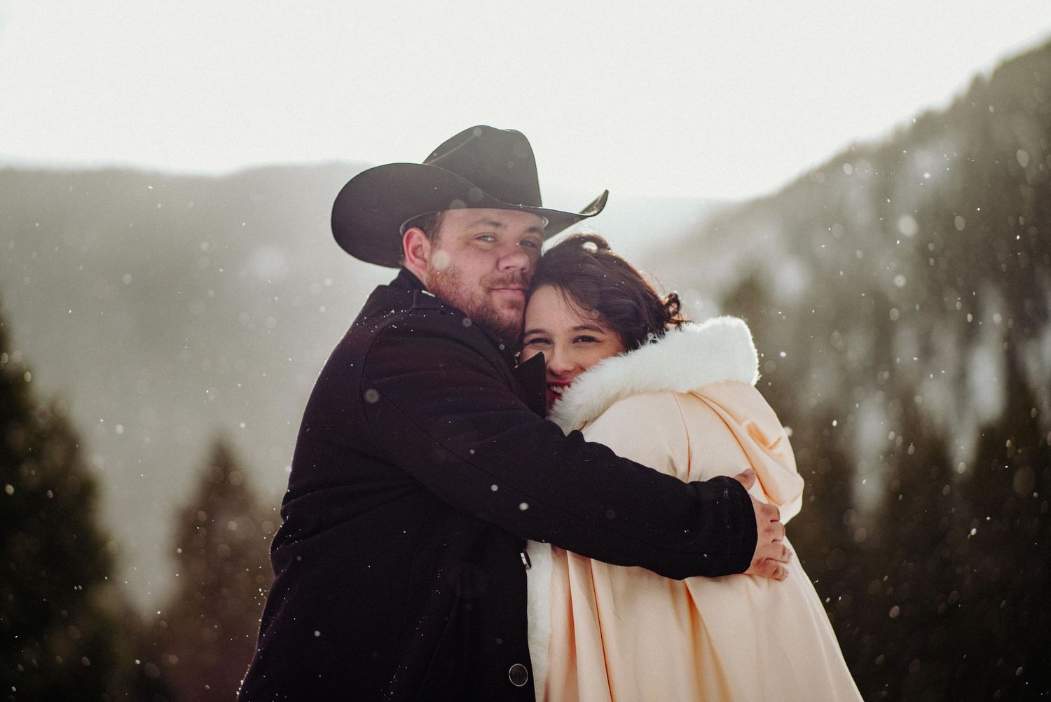 Couple embracing in snowy mountains, man in black cowboy hat hugging woman in white coat, romantic winter elopement.