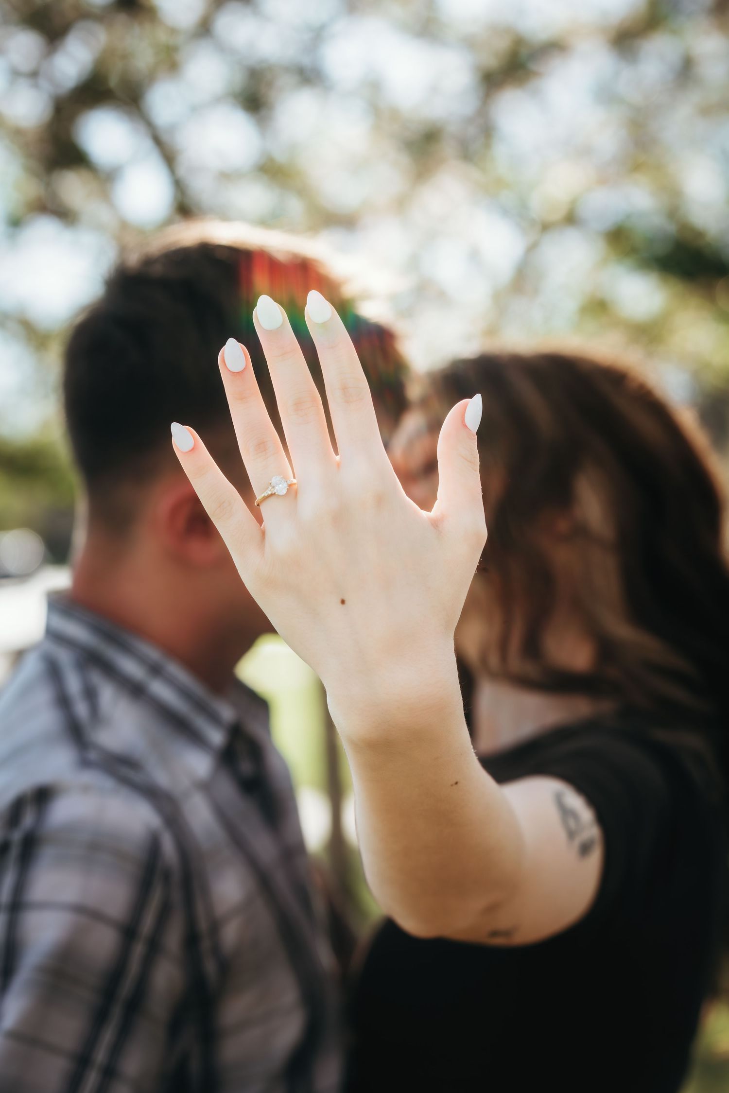 Woman shows off engagement ring while couple kisses in bokeh park background.