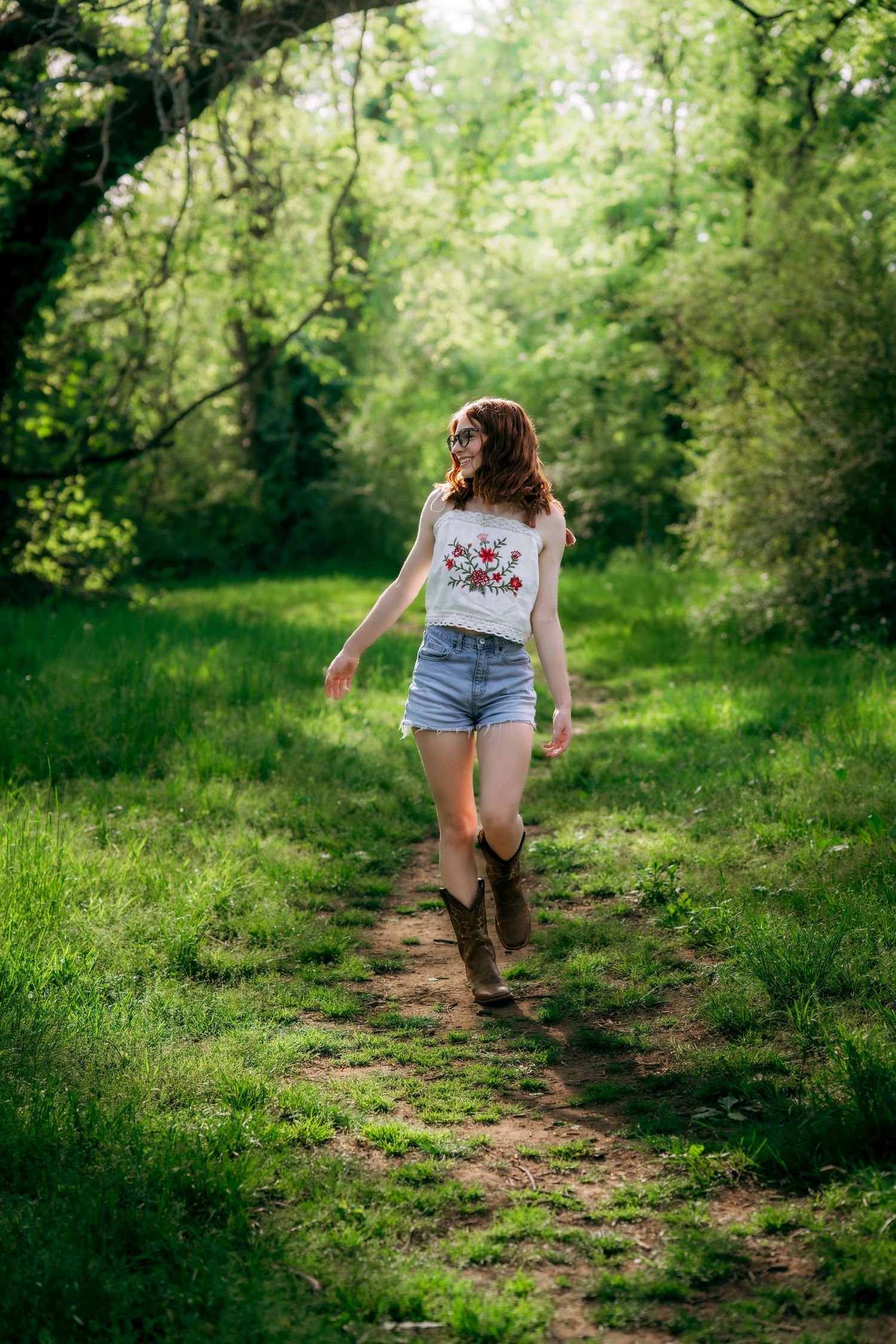 Young woman in floral top, denim shorts, and cowboy boots walking along a lush green forest path.