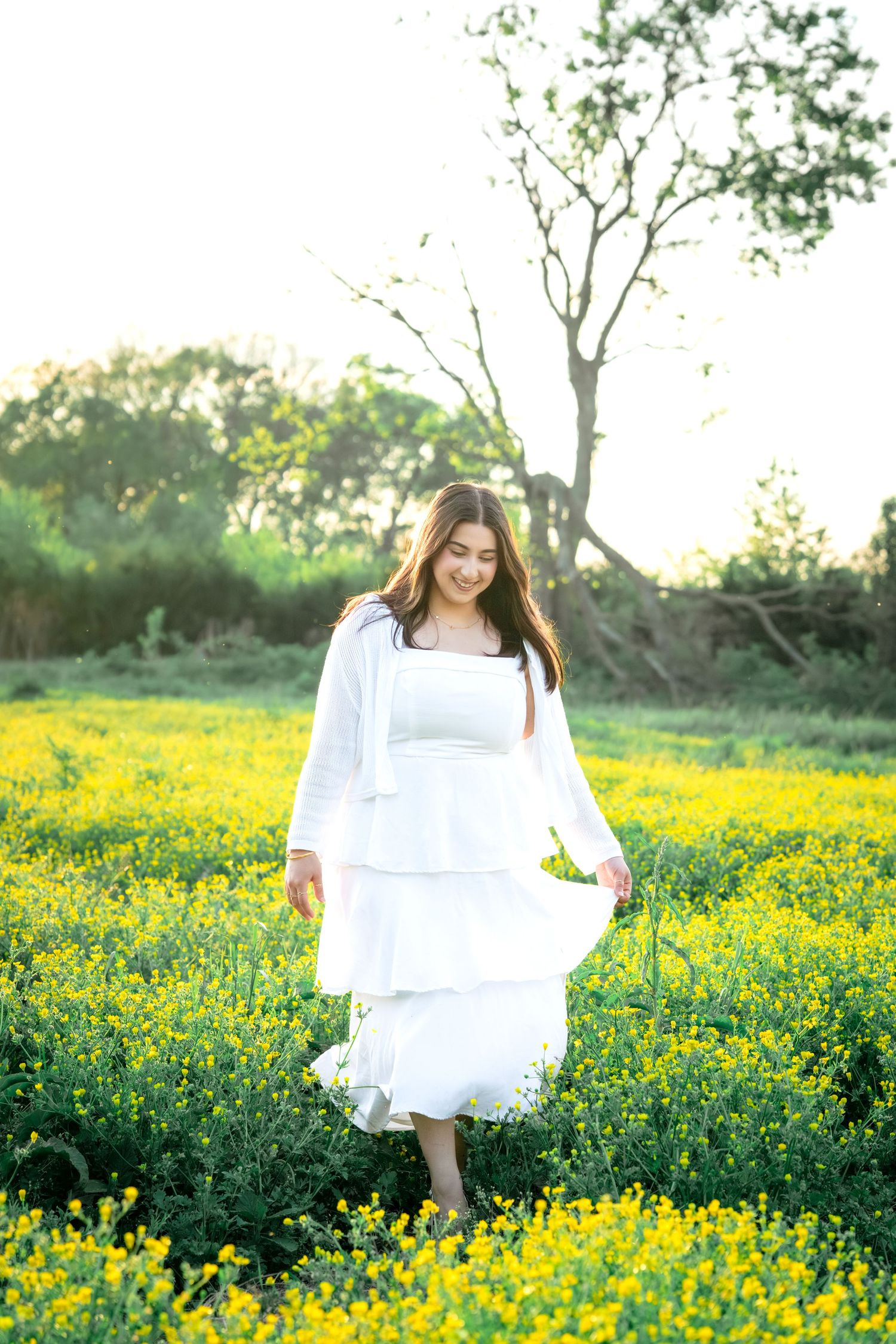 Young woman in white tiered dress walking through vibrant yellow wildflower field at sunset.