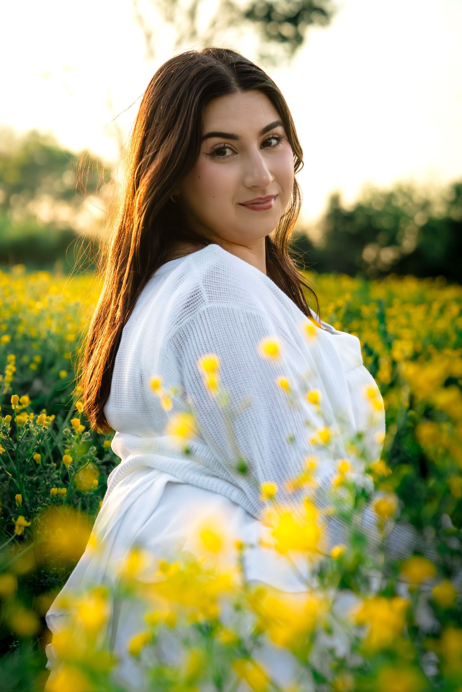 Young woman in white dress smiling in a vibrant yellow flower field during golden hour sunset.