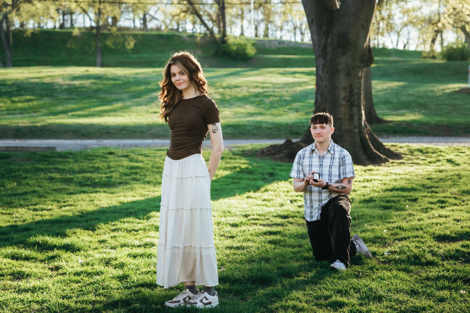 A man kneels on green grass proposing to a woman in a white skirt near a large tree in a sunny park.