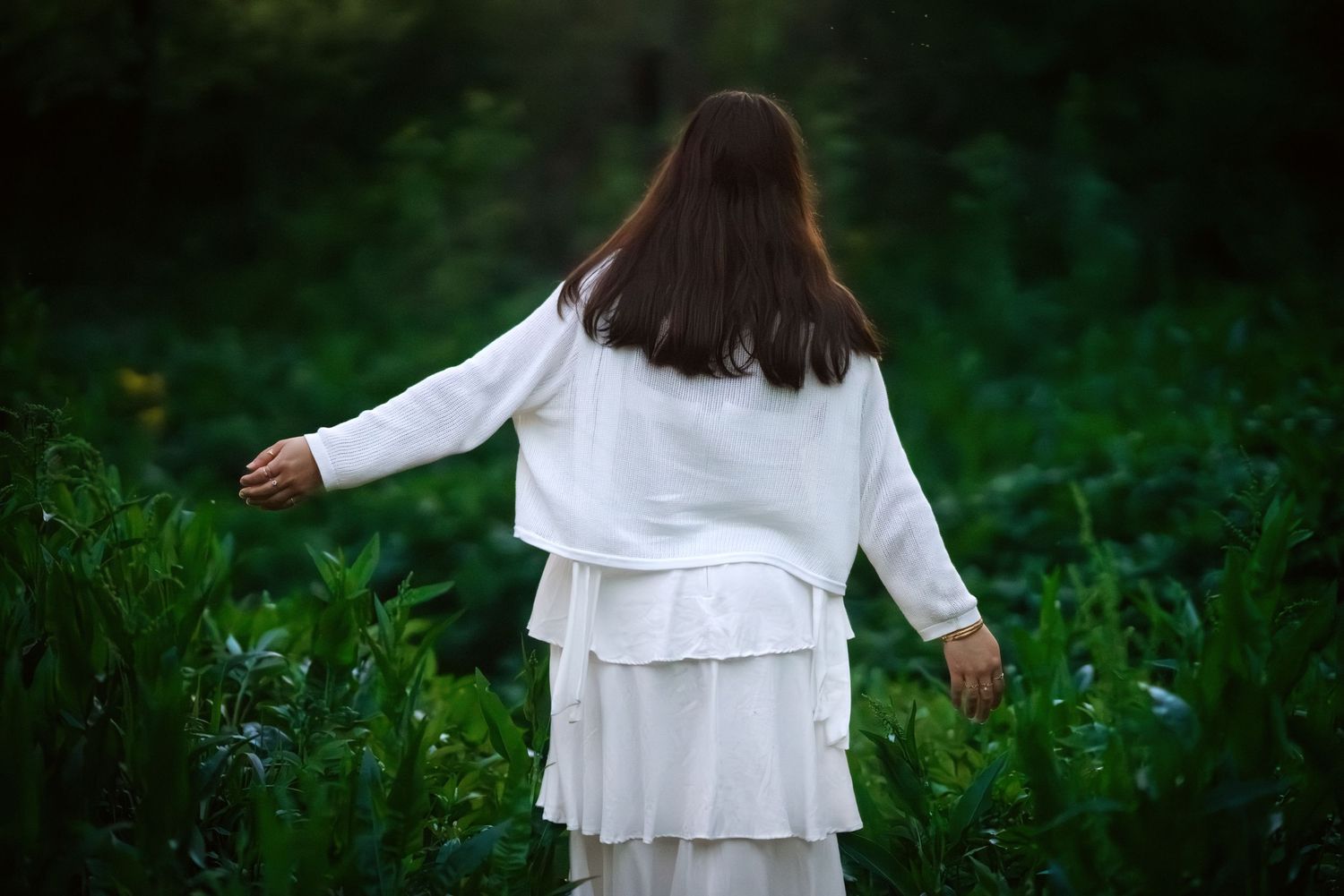 Woman in white dress walking through lush green field, viewed from behind with arms outstretched.