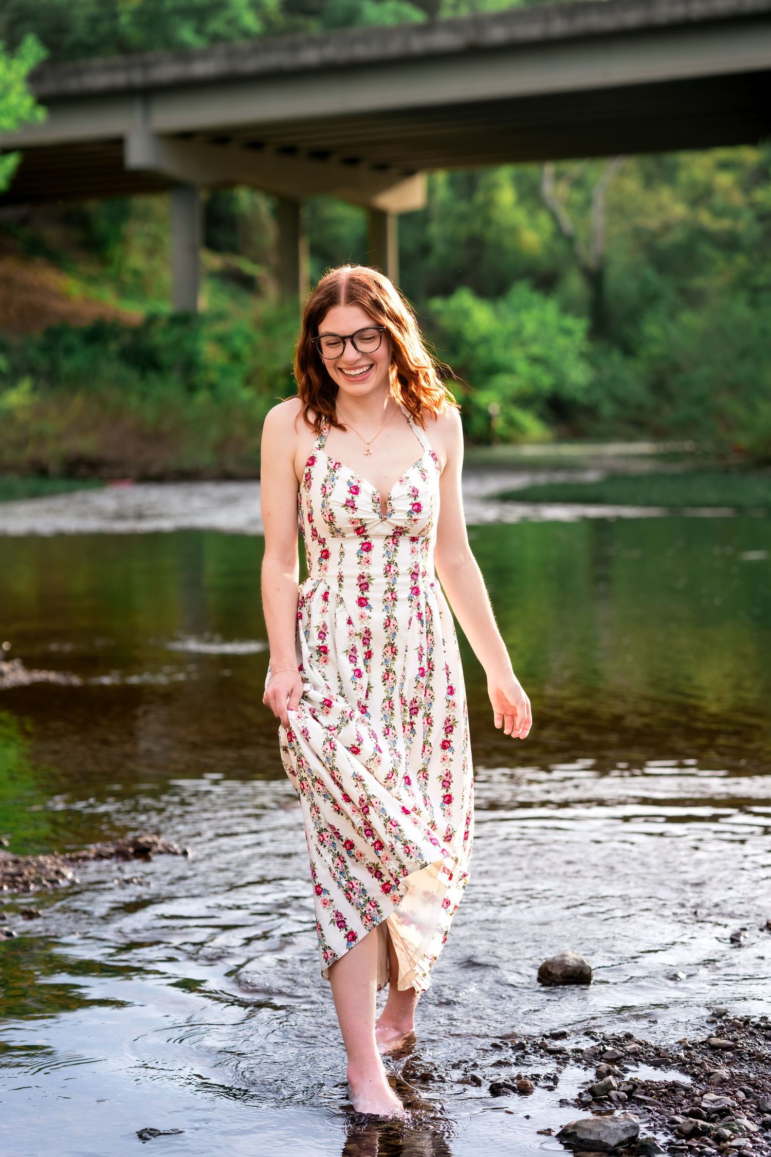 Young woman in floral sundress and glasses wading barefoot through a shallow creek under a bridge.