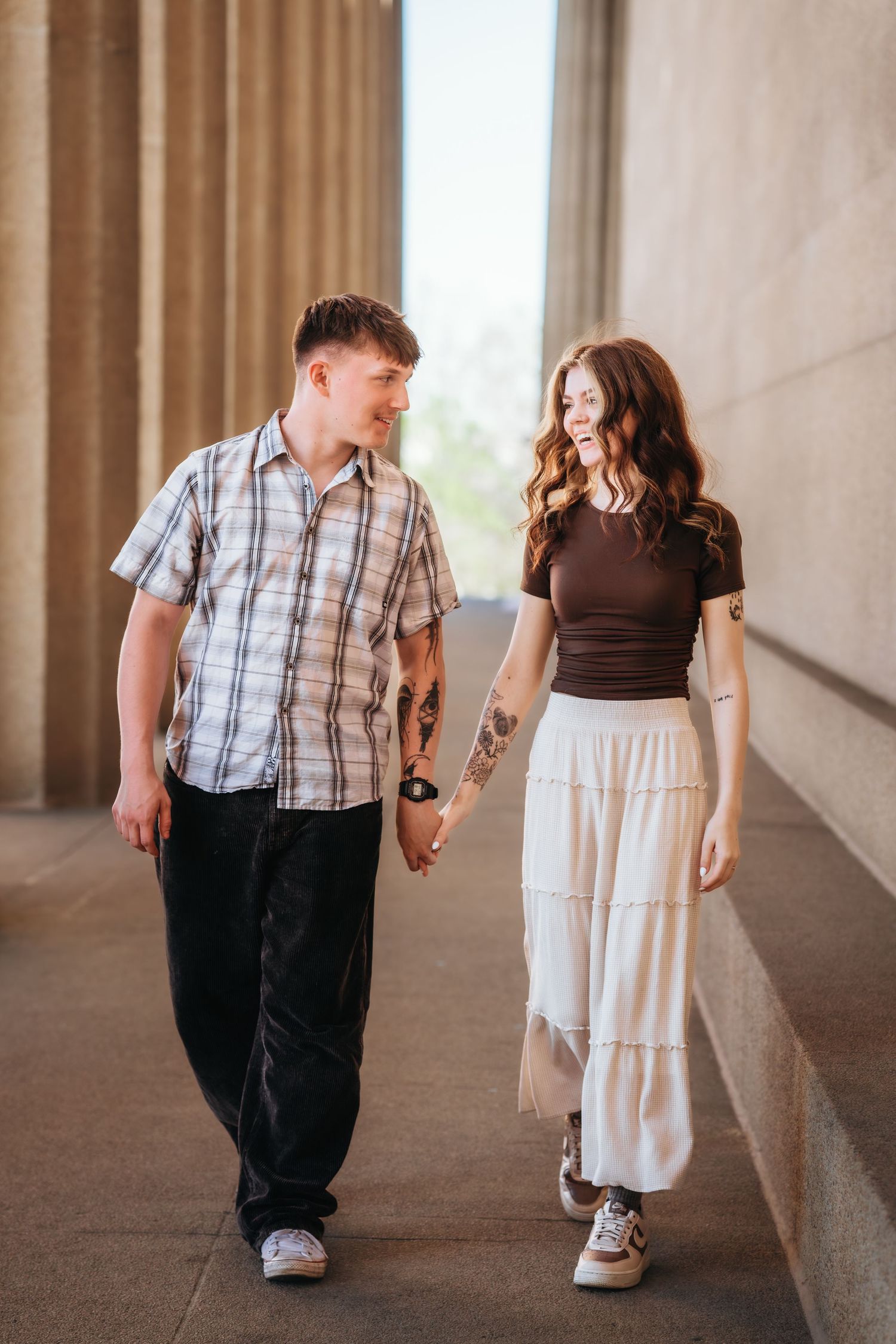 A tattooed couple holds hands and walks together through a columned outdoor corridor, smiling at each other.