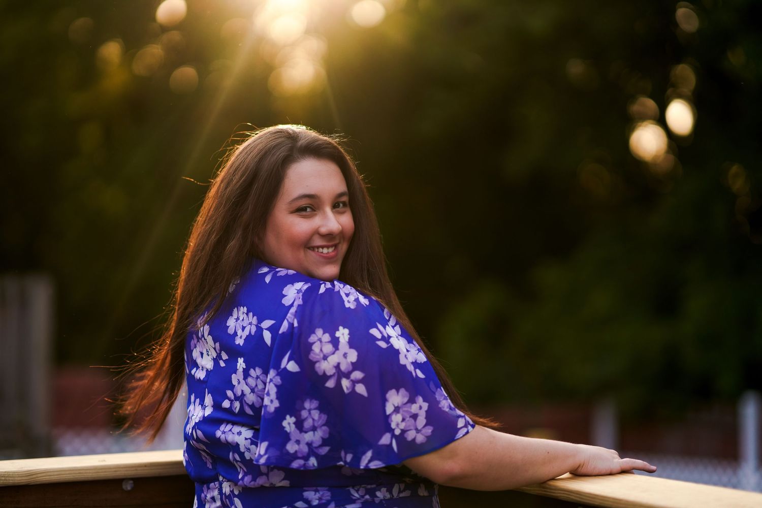 Woman in blue floral dress smiling over shoulder at golden hour sunset outdoors.