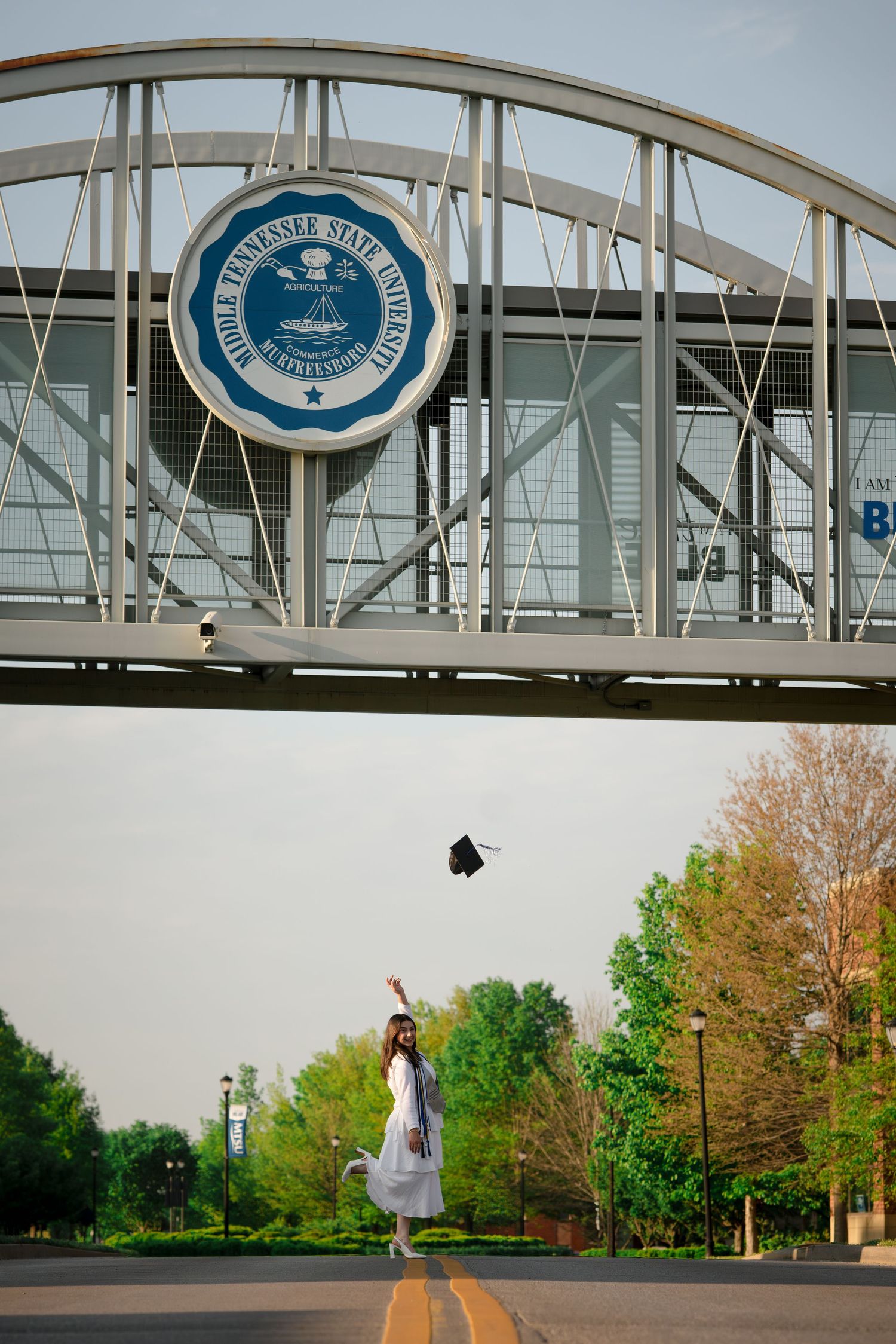 Graduate tosses cap in air beneath Middle Tennessee State University bridge sign on a tree-lined campus road.