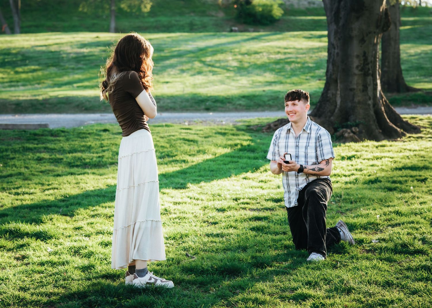 Man kneeling on one knee proposing to woman in white skirt on green grass park lawn in golden sunlight.