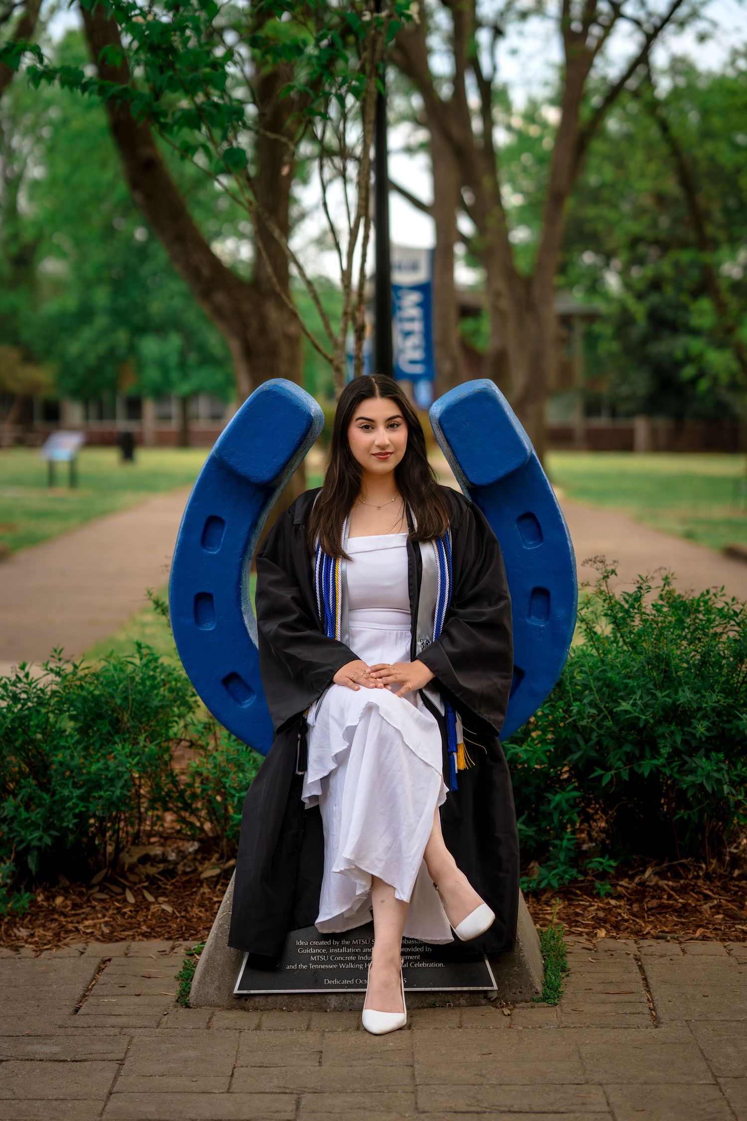 Young woman in graduation gown poses in front of blue horseshoe statue at MTSU campus.