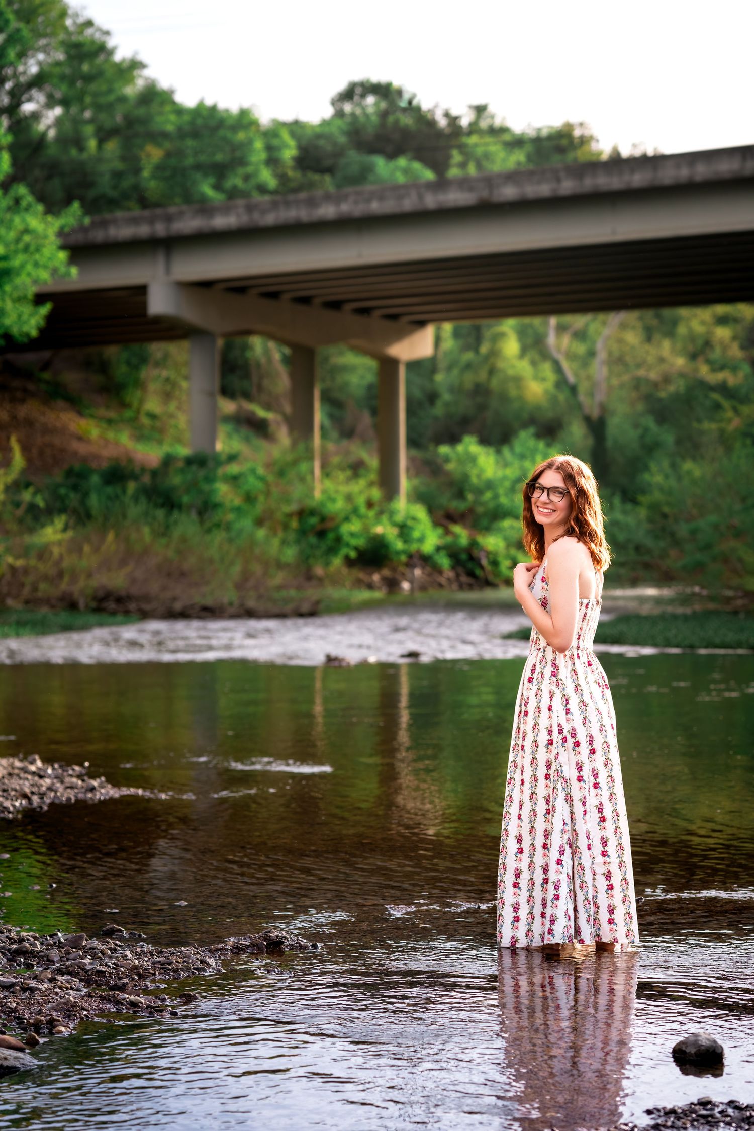 Young woman in floral maxi dress standing in a shallow river with a bridge in the background.