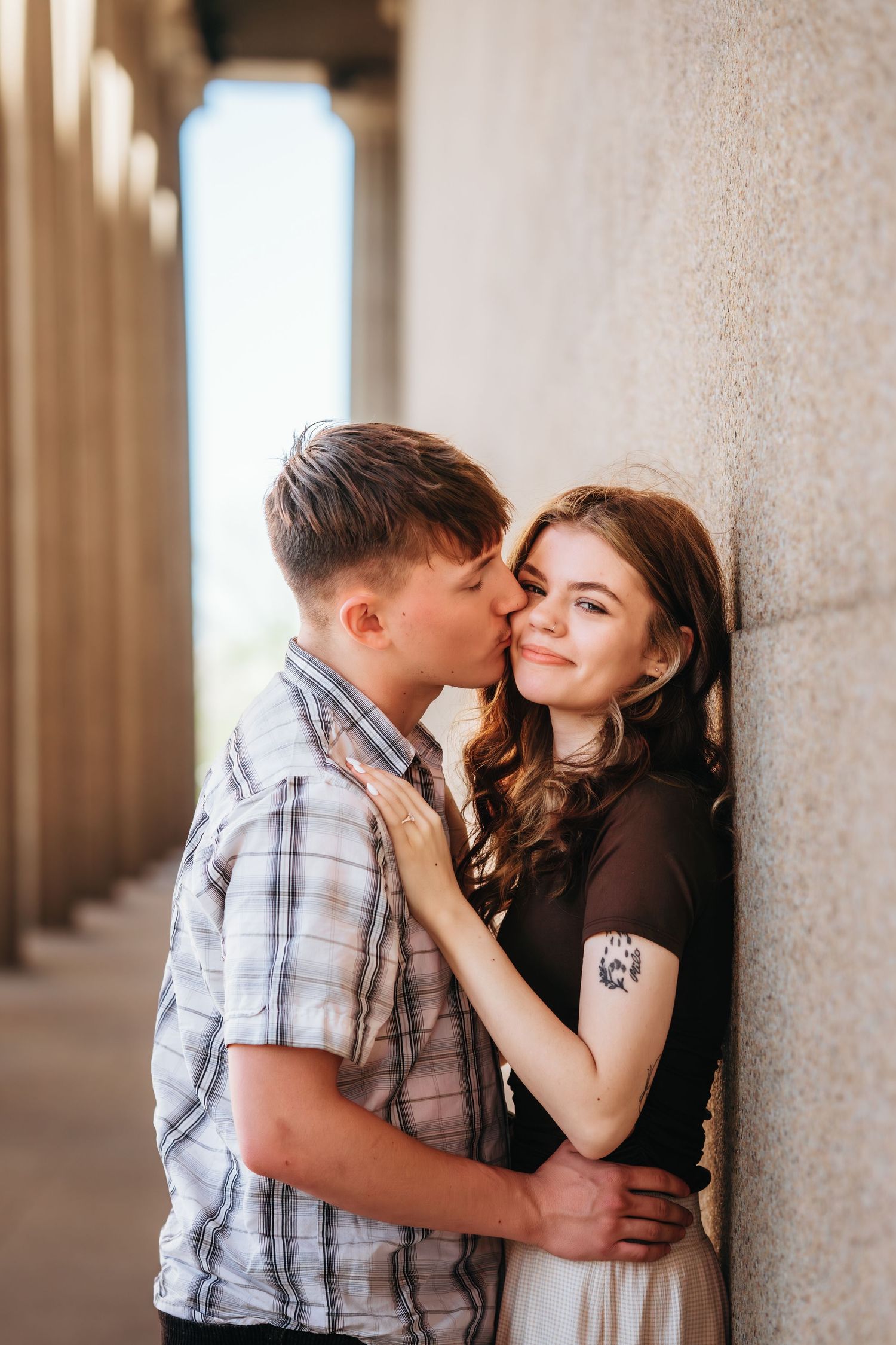 A young man kisses a smiling woman on the cheek as she leans against a textured stone wall amid tall columns.