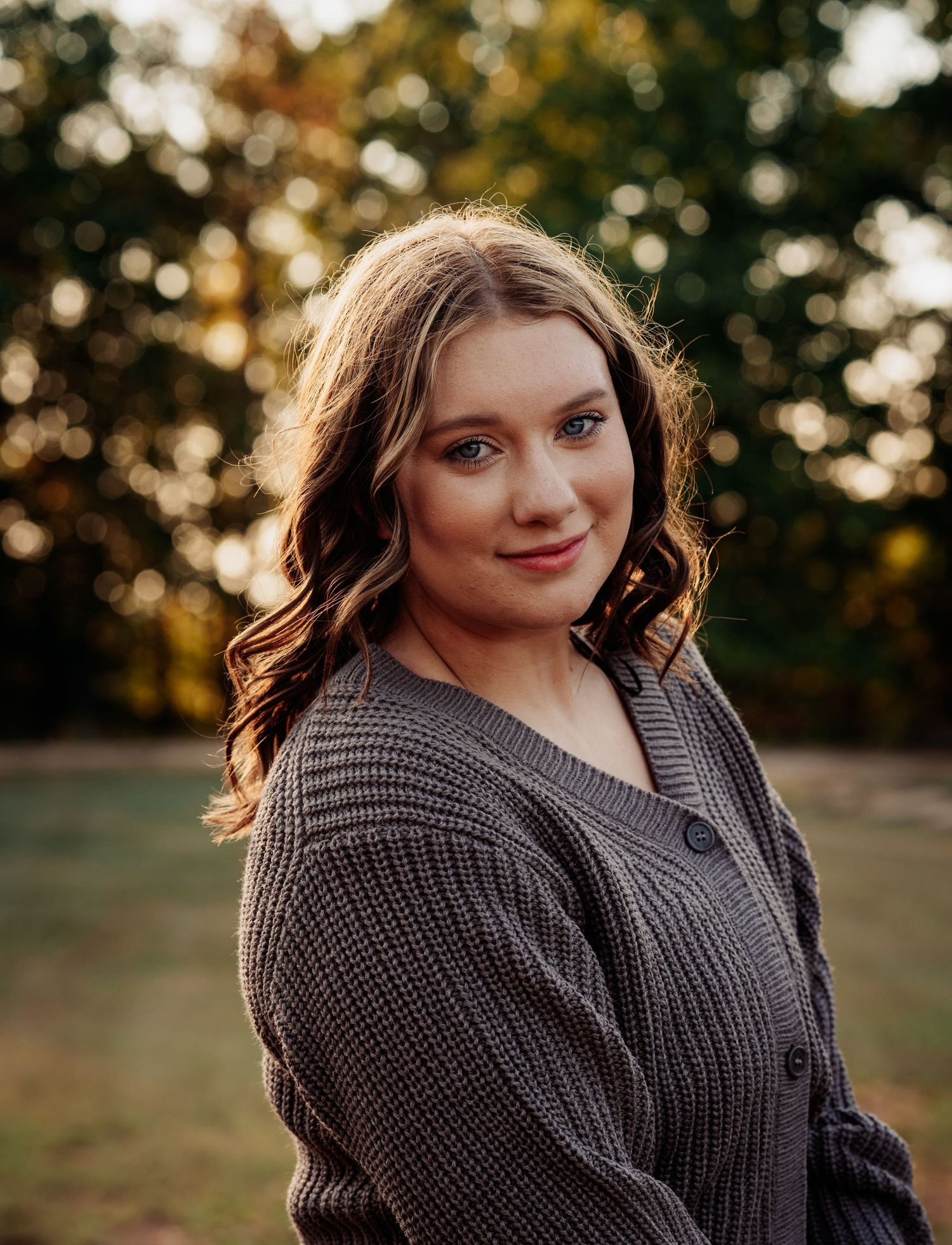 Young woman in gray knit cardigan smiling outdoors with bokeh autumn trees background in golden hour light.