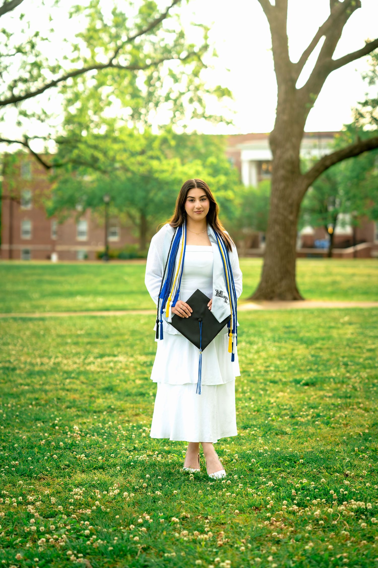Young woman in white dress holding graduation cap on green campus lawn with trees and brick building.
