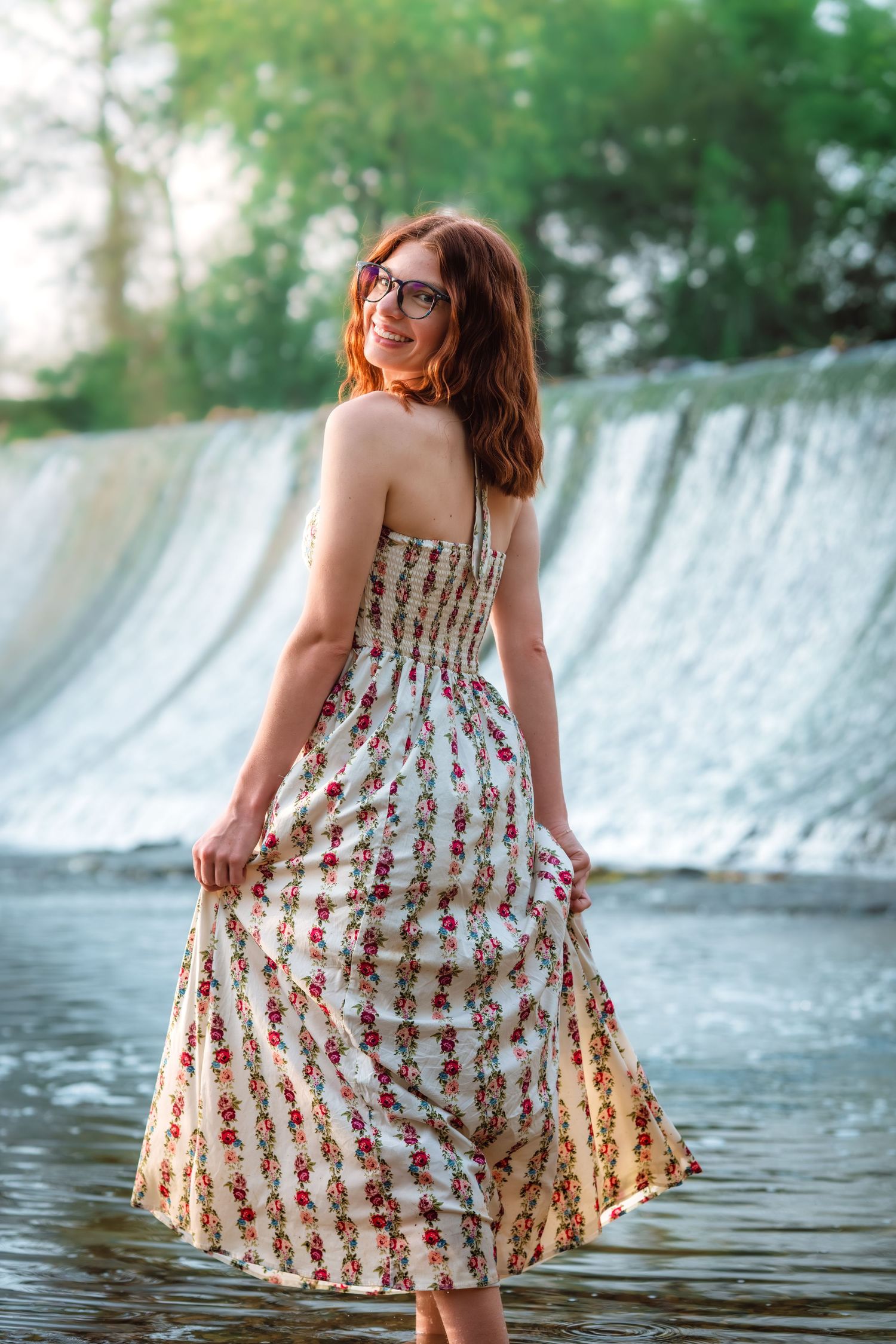 Redhead woman in floral sundress standing in shallow water near a waterfall in a lush forest.