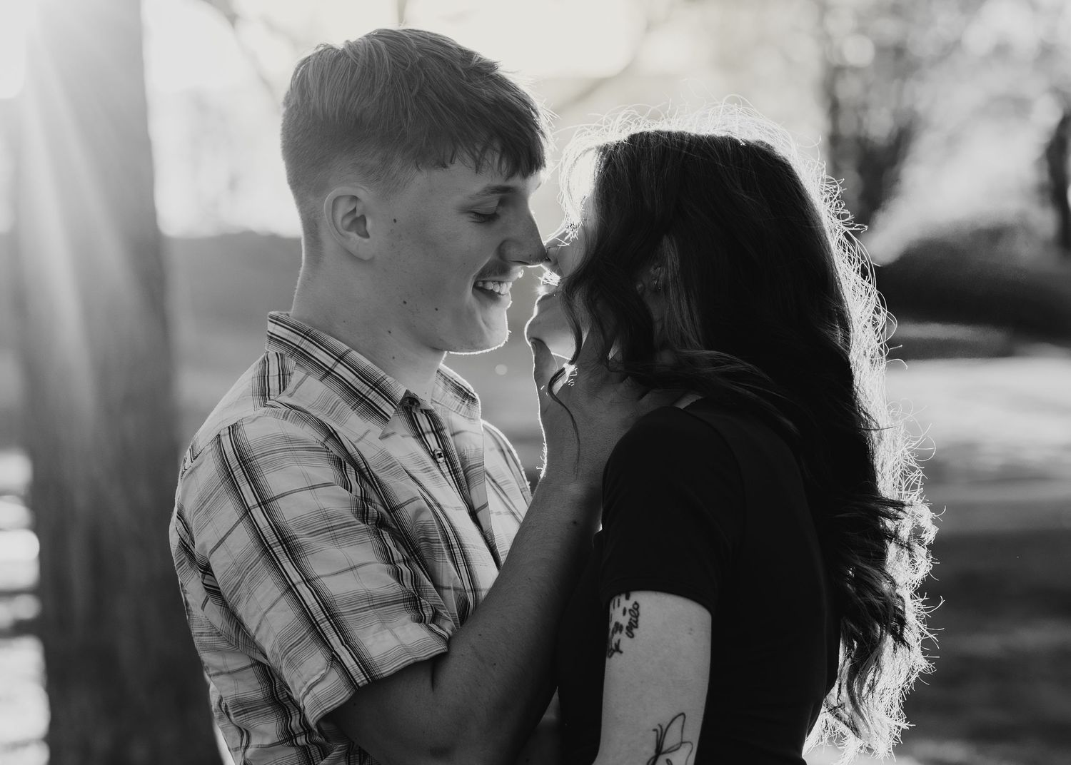 Black and white photo of a smiling young couple sharing a romantic moment outdoors in soft sunlight.