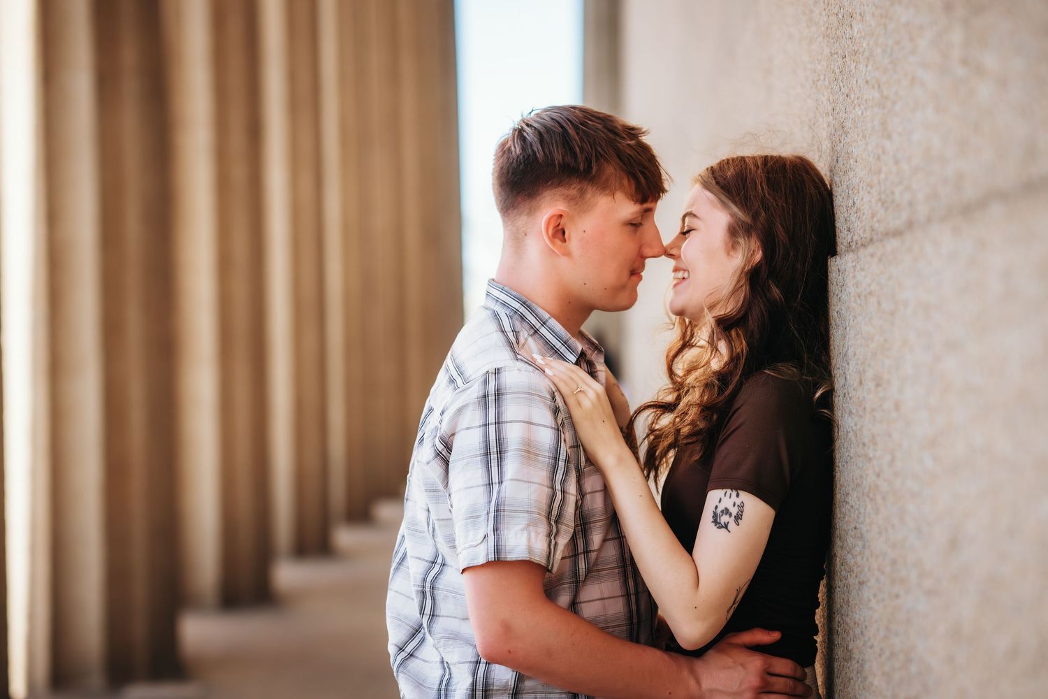 A young couple shares a romantic moment, nearly kissing while leaning against a stone wall between tall columns.