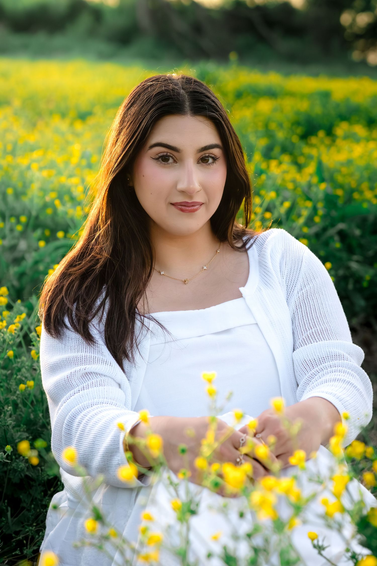 Beautiful young woman in white outfit sits among vibrant yellow wildflowers in golden hour light.