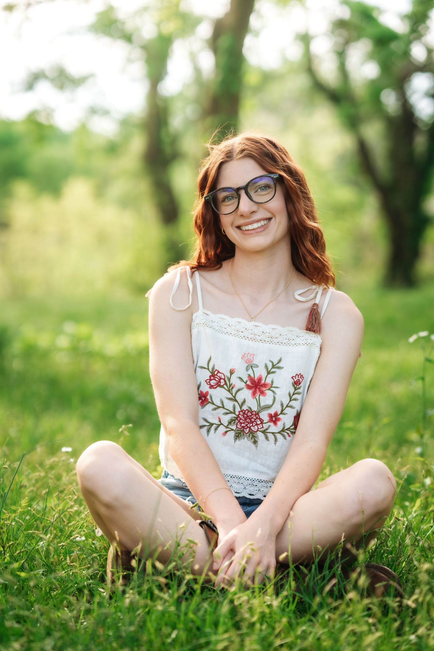 Smiling redhead woman with glasses sitting cross-legged on green grass wearing floral embroidered top and denim shorts.