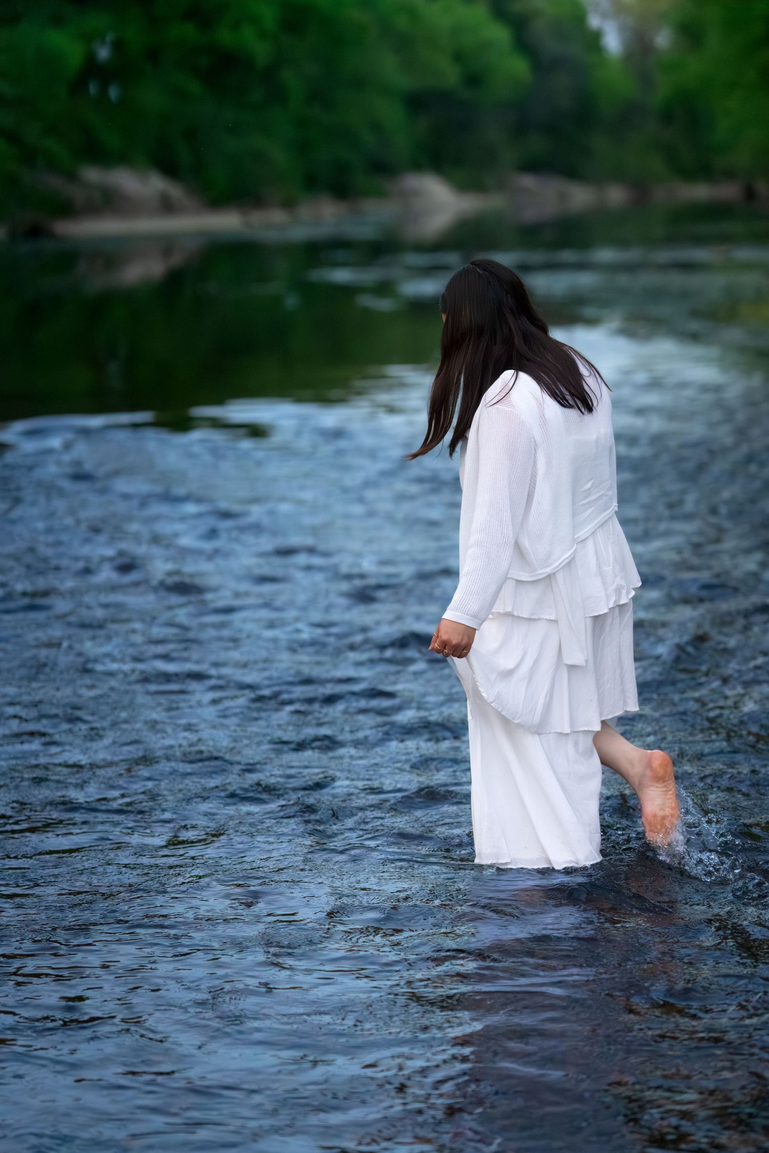 Young woman in flowing white tiered dress wades barefoot through a calm shallow river outdoors.