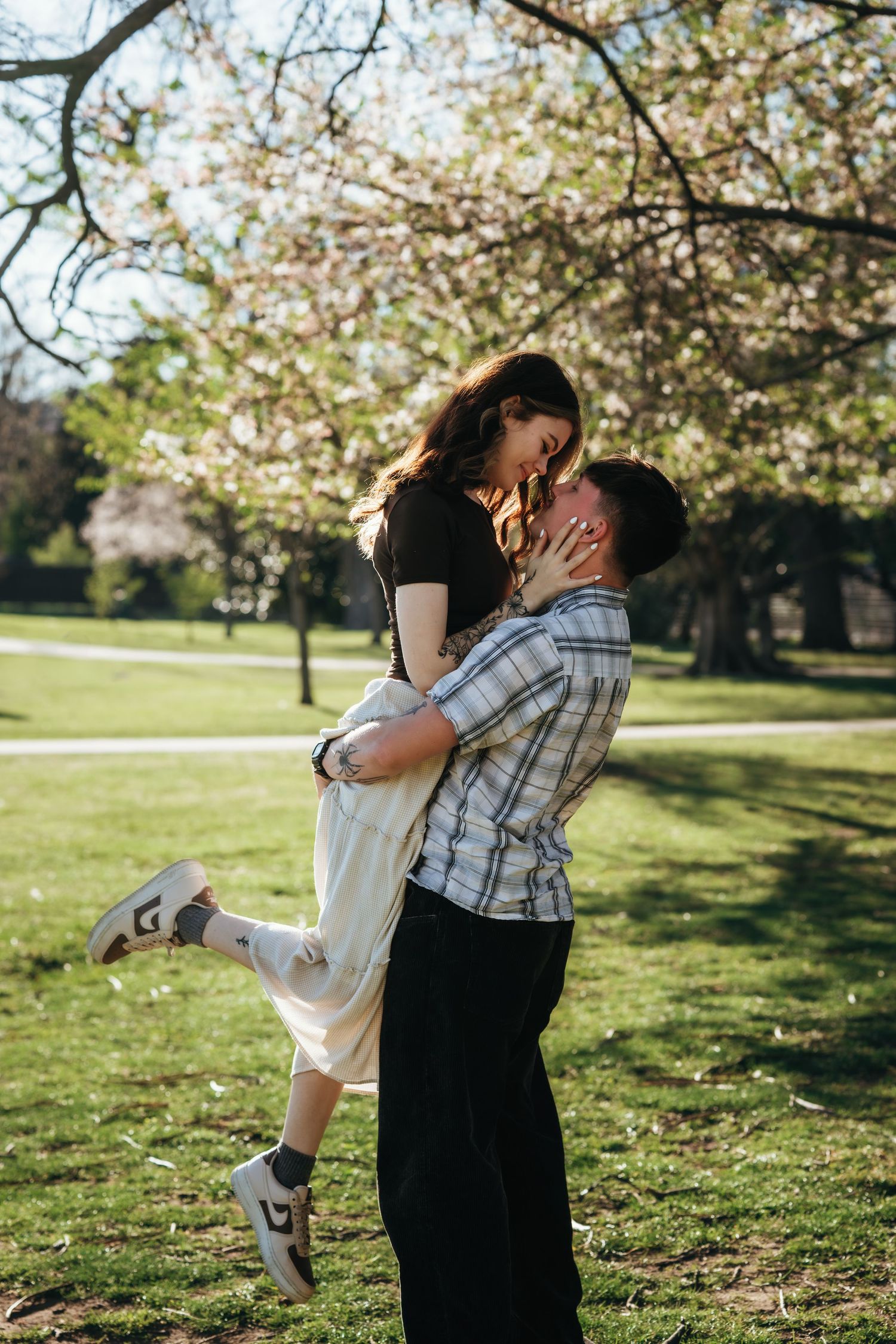 Man lifting woman in a joyful embrace outdoors in a sunlit park with blooming trees in the background.