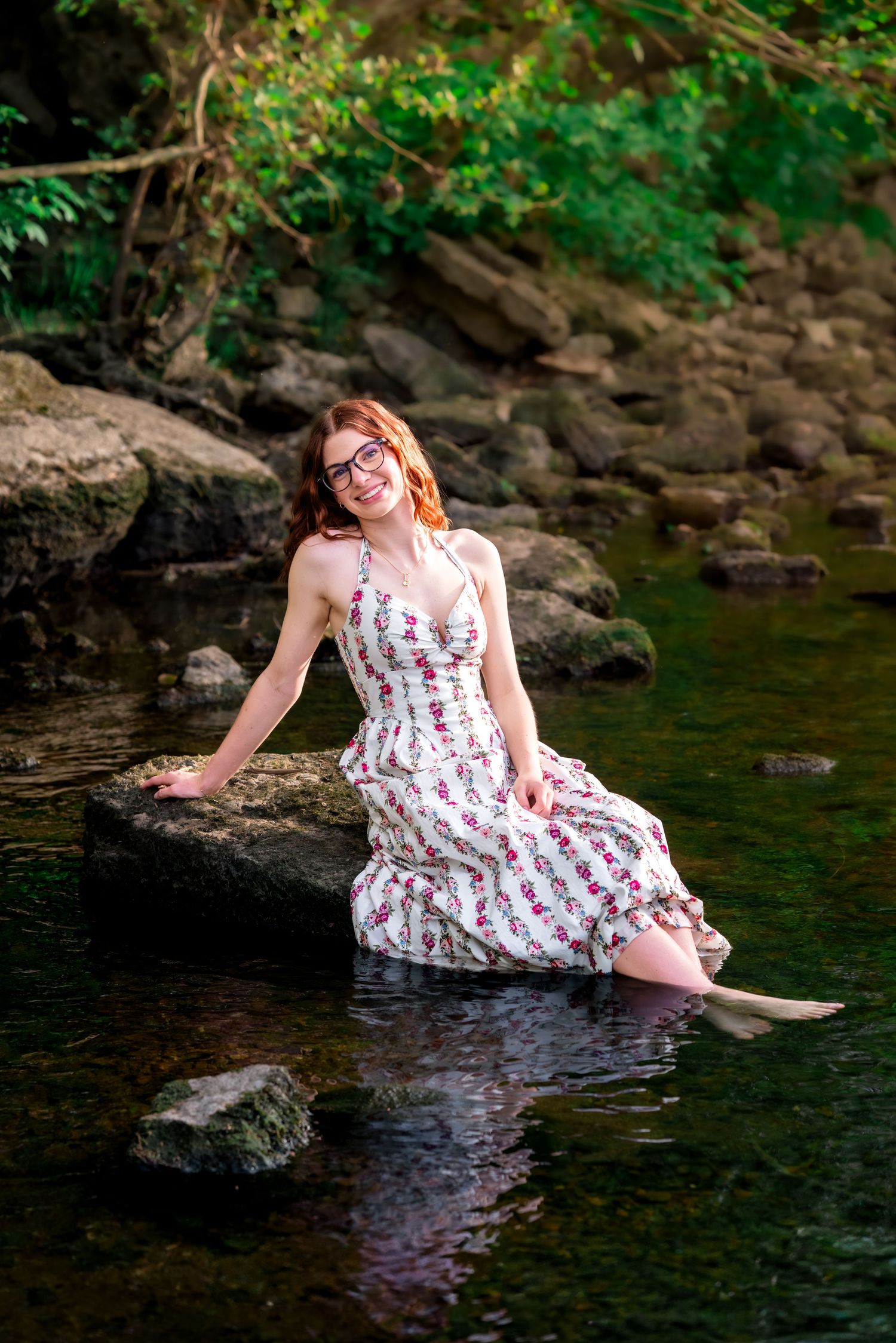 Young redhead woman in floral sundress sitting on rocks in a scenic woodland creek.
