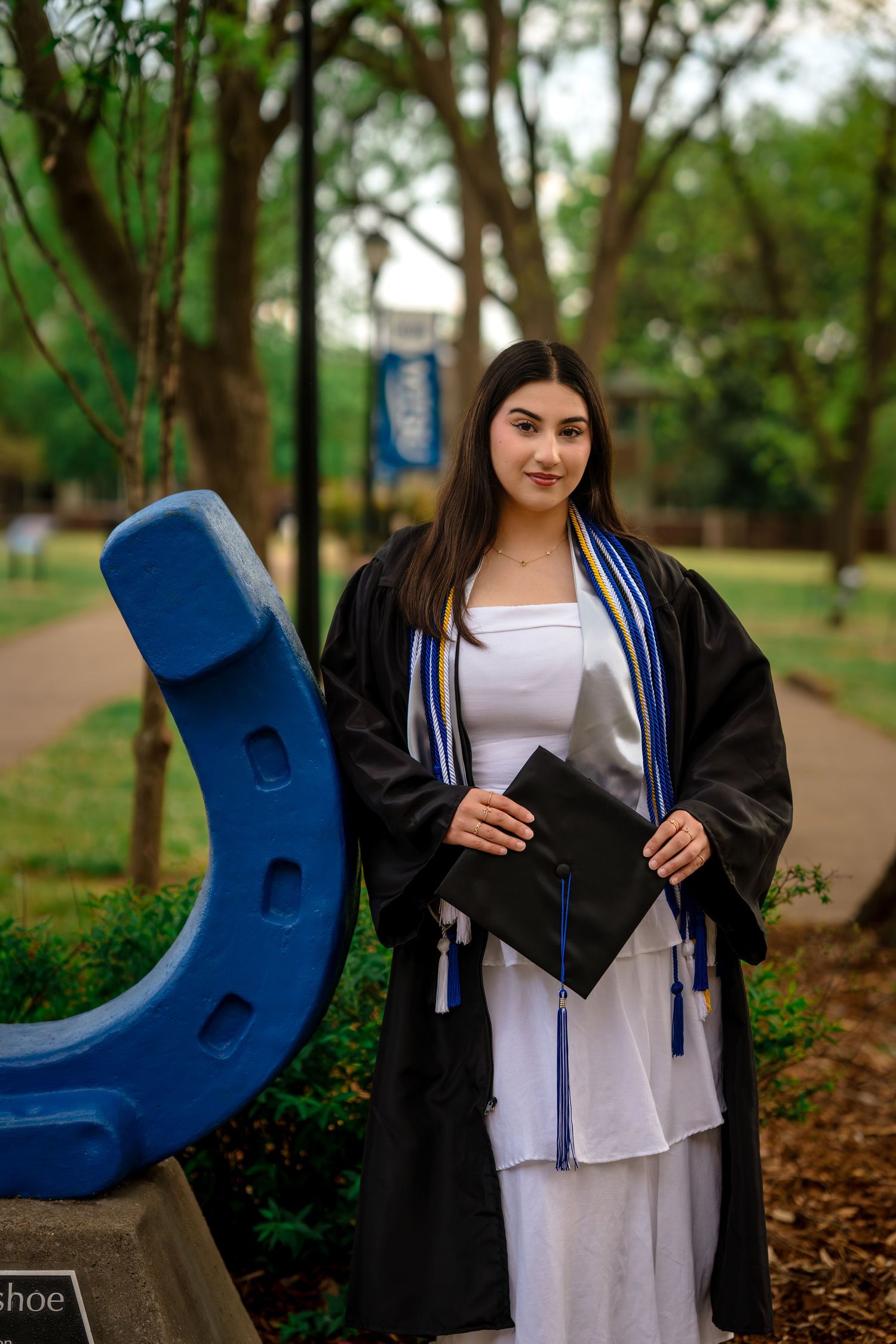 Young woman in graduation gown and white dress poses beside blue horseshoe sculpture on college campus.