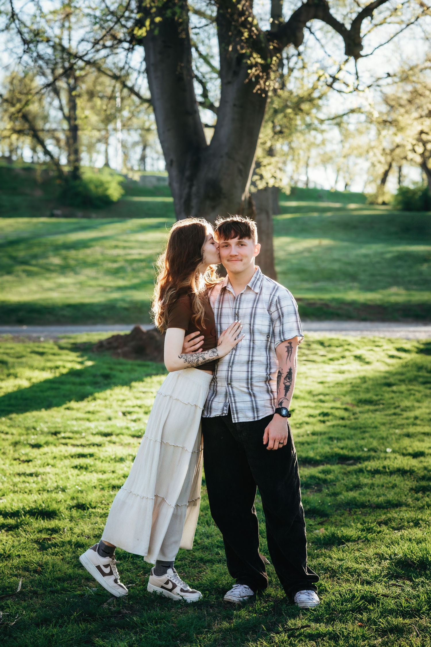 Couple posing in a lush green park, woman kissing man on the cheek beside a large tree in warm sunlight.