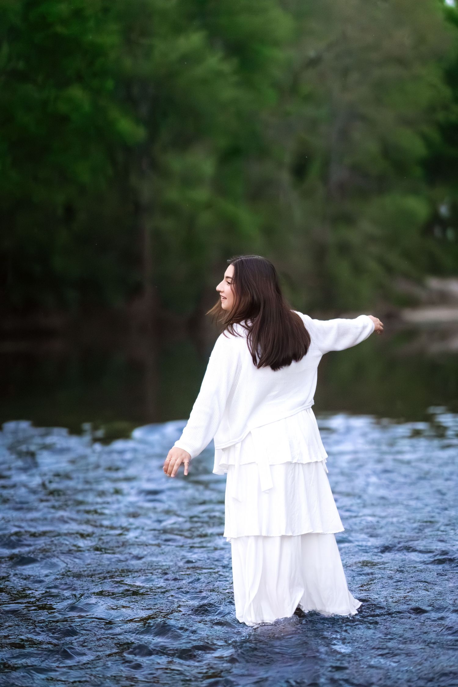 Woman in white dress wading through a serene river surrounded by lush green trees.