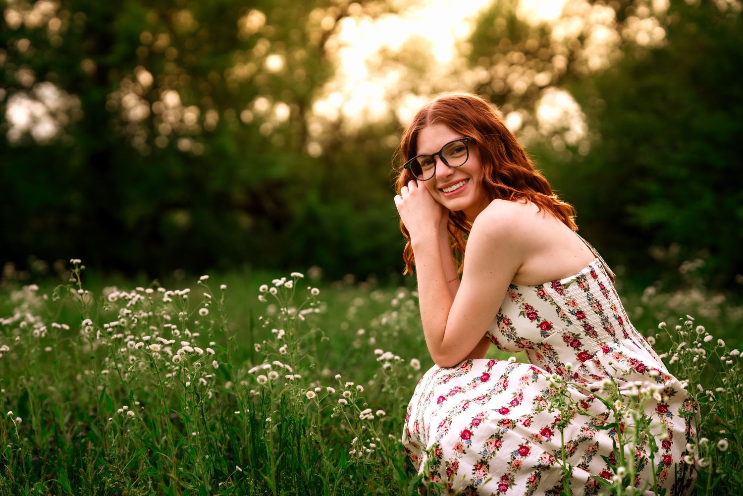 Young redhead woman in floral dress and glasses smiling in a wildflower meadow at golden hour.