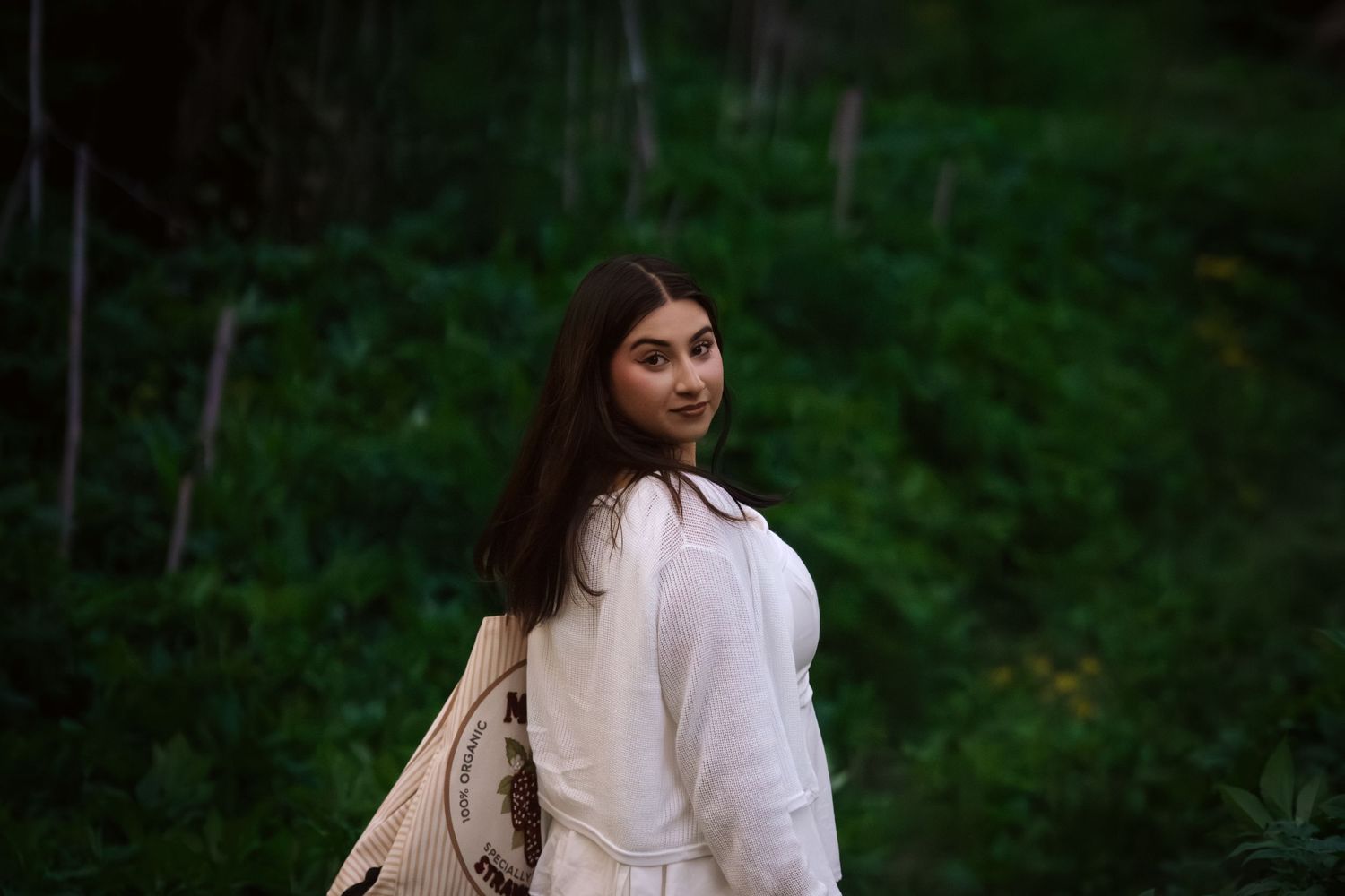 Young woman in white blouse with tote bag glances back over shoulder in lush green forest setting.