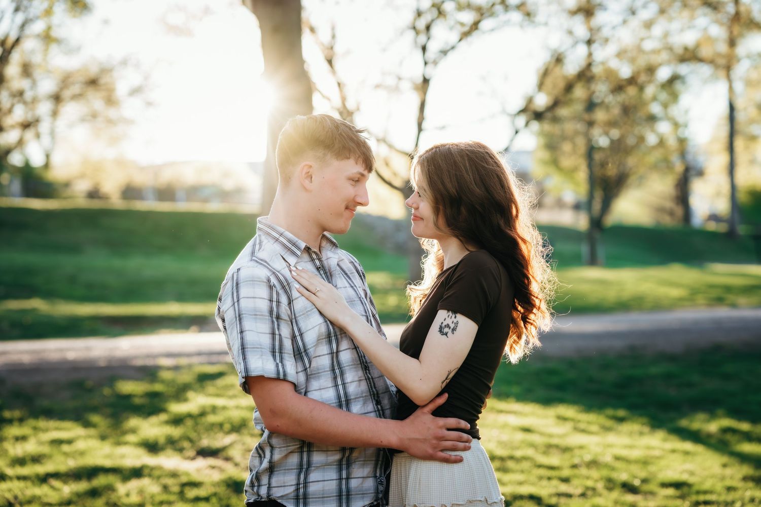 Couple embracing and smiling at each other in a sunny park with green trees in the background.