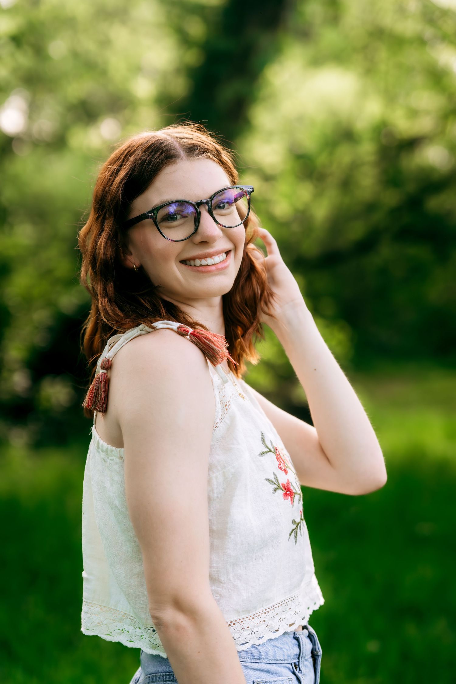 Smiling young woman with red hair and glasses wearing a white floral embroidered top outdoors in a green park.